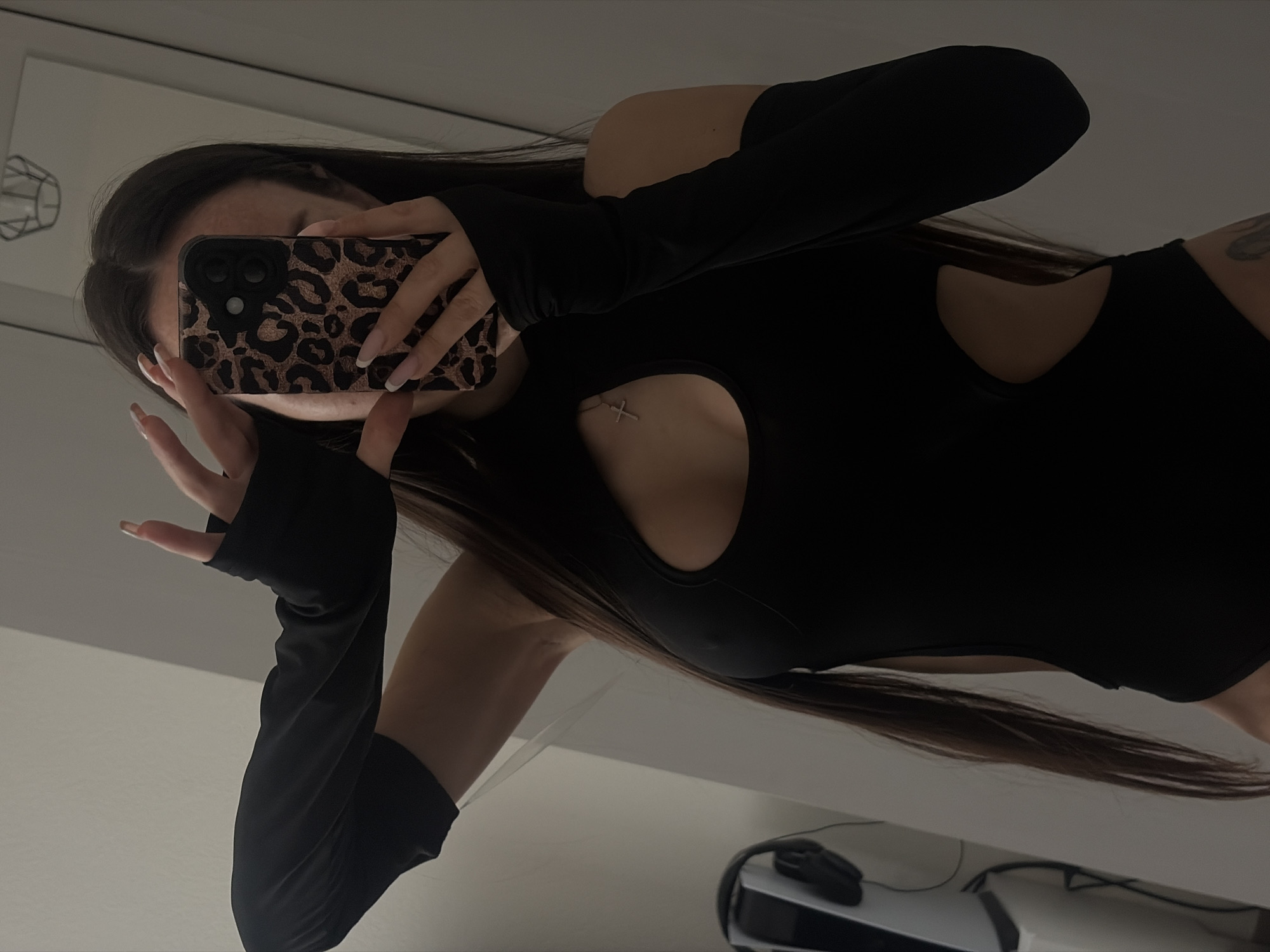 A woman in a black, long-sleeved, cut-out bodysuit takes a mirror selfie. The phone she is holding has a leopard print case. A glimpse of a silver cross necklace is visible above the chest cutout. The background suggests an indoor environment. No information indicates a specific location in Saint Petersburg, Russia.