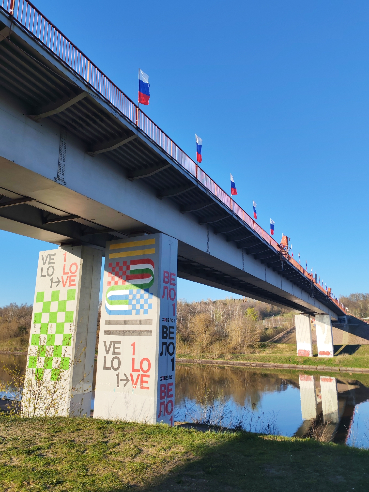 The image captures a bridge spanning a river, likely in Yakhroma, Russia, as the Russian flags lining the bridge's railing suggest. The bridge's structure is predominantly gray, with a red railing along the top. The supports of the bridge are painted with colorful designs and text, including the word "VELO" and the phrase "1 LOVE". The surrounding environment consists of green grassy areas and trees in the background. The river's surface reflects the bridge supports, creating a mirrored effect. The sky is a clear, bright blue, suggesting a sunny day.