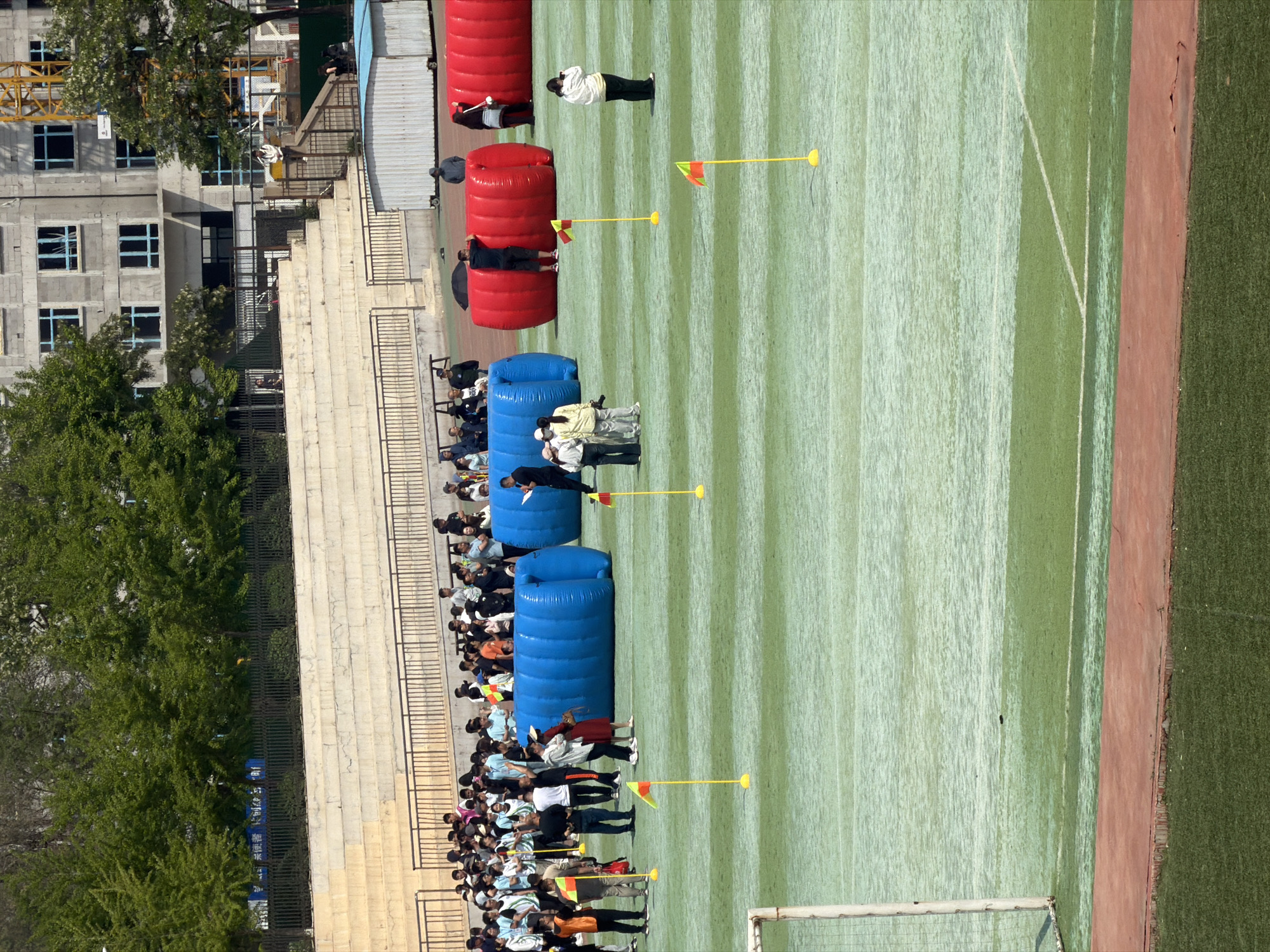 A high-angle shot depicts an outdoor paintball field setup, presumably in Zhongtun, China. The field, marked with parallel light and dark green stripes, is flanked by a dirt track on the right and stadium seating on the left. A crowd of people, seemingly spectators, lines the seating area. Along the field, inflatable paintball bunkers alternate in blue and red. Individuals in casual clothing stand between the bunkers, some appearing to be referees with yellow flag markers nearby. A goalpost is visible in the lower left corner. In the background, a building under construction with a crane is visible. Trees are on the left.