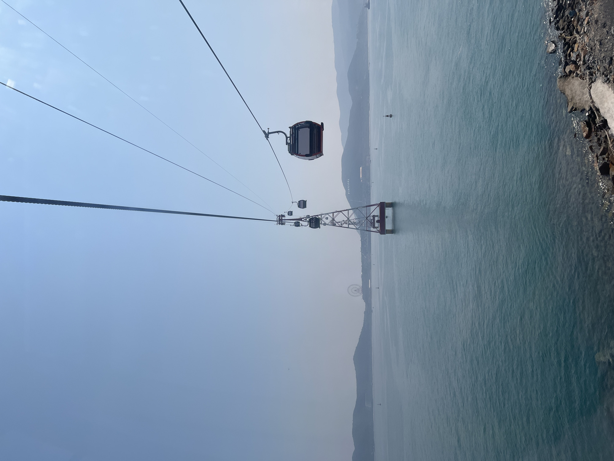The image shows a cable car traversing the sea towards a support tower, with a coastline visible in the background. The tower appears to be anchored in the water. The location is in Nha Trang, Vietnam. The sky is clear and the water is calm. A rocky coastline is visible on the right side.
