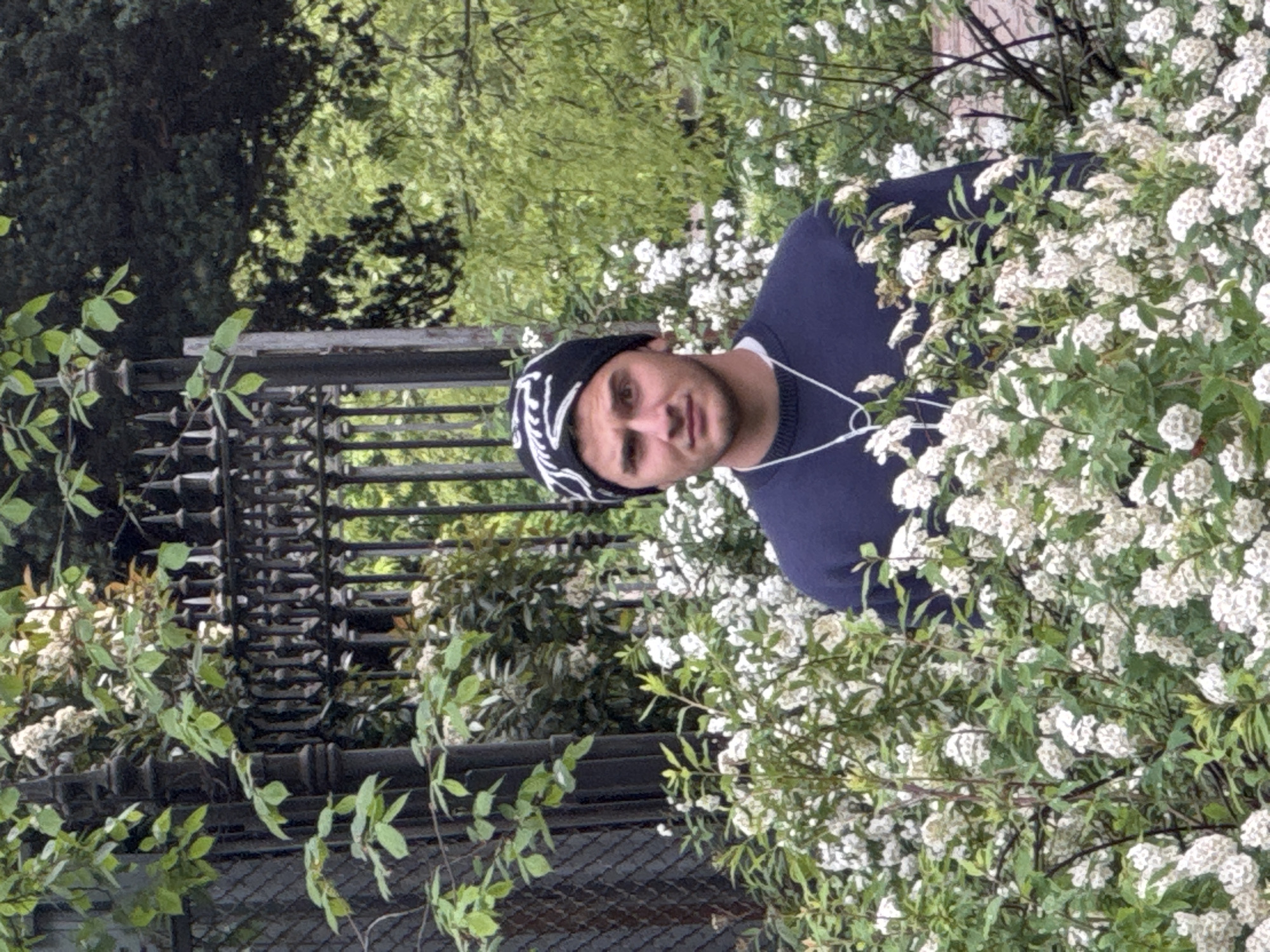 A man wearing a dark blue sweater and a black beanie with white patterns stands in front of a metal fence, partially obscured by flowering bushes with small white blossoms. The location appears to be in a park or garden, given the presence of greenery and trees in the background. Based on the architecture of the fence, it is plausible that the location is in Milan, Italy.