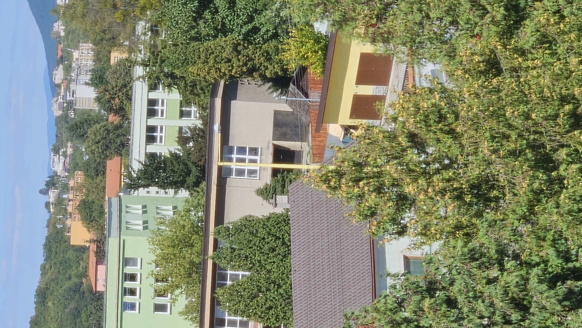 The image shows an aerial view of a residential area in Prešov, Slovakia. In the foreground, there are several trees, some of which are deciduous and showing autumn colors. Behind the trees, there are multiple buildings. The closest building has a brown, tiled roof and light-colored walls. Adjacent to it is a yellow building with two brown garage doors. Further in the background, a long, light-gray building with multiple windows is visible, and behind that, a multi-story green building with numerous windows. In the distance, the landscape rises into a hill covered with trees, and beyond that, more buildings and houses are scattered across the slopes, with mountains visible in the far distance under a clear blue sky. The scene appears to be a typical urban or suburban neighborhood during the daytime.