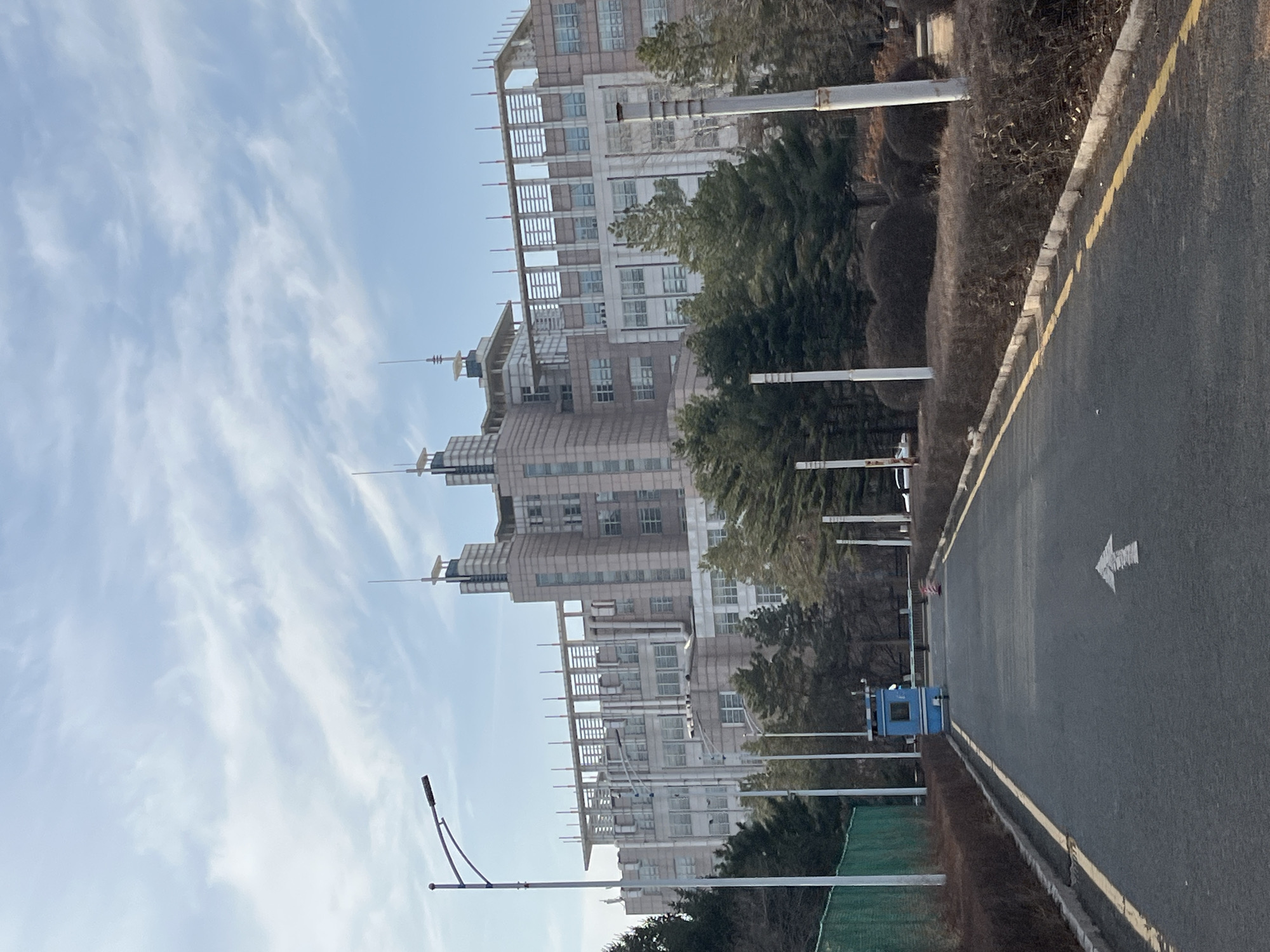 The image depicts a multi-story building complex with a modern architectural style, likely located in Changchun City, China. The buildings are predominantly beige or light gray, with numerous windows and distinctive rooftop structures that resemble antennae. A road runs alongside the buildings, featuring painted directional arrows. The road is edged with a curb and bordered by shrubs and trees. The sky is light blue with scattered clouds. A lamp post is visible near the road. A blue electrical box and green netting appear along the road. There are no visible people.