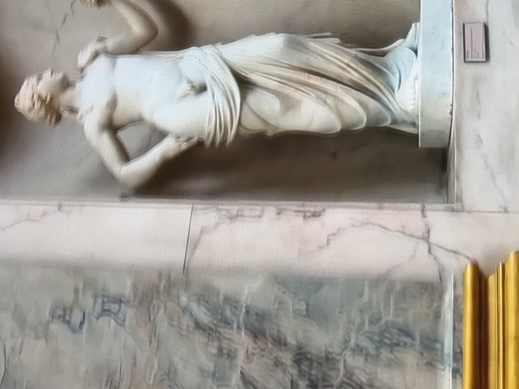 The image shows a marble statue of a person with androgynous features, draped in a toga-like garment. The statue is white and stands on a white pedestal. To the left of the statue, there is a marble wall with veining. In the foreground, a golden wooden ledge is visible. A small plaque with text is placed on the white surface below the pedestal, to the right. The lighting in the image is soft and diffused. The statue appears to be located indoors, possibly in a museum or gallery. The background is a neutral, light-colored wall, and the overall environment suggests a display or exhibition setting.