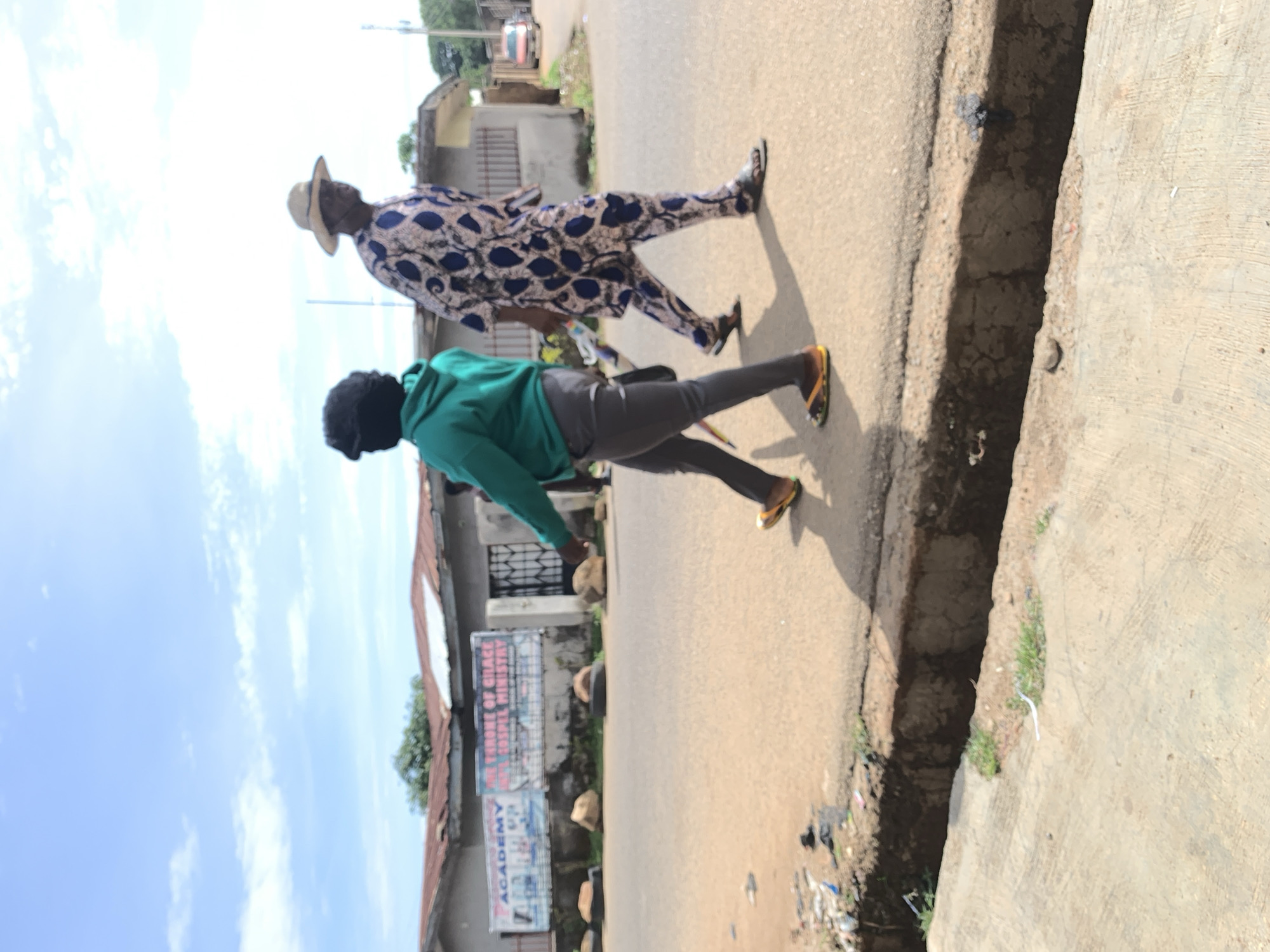 This image captures a street scene in the city of Jos, Nigeria, under a bright, partly cloudy sky. Two individuals are walking from left to right on a light-colored asphalt road. The lead person wears a green hoodie, dark grey pants, and multi-colored sandals; their head is obscured by a dark covering, and a patterned object is partially visible in their left hand. Trailing slightly, the second person wears a patterned blue and light-colored tunic with matching trousers, sandals, and a straw hat. Both cast distinct shadows on the road, indicating bright overhead sunlight.
To the immediate left of the road, an open drainage ditch with exposed earth and concrete runs parallel, separating the road from a lower concrete pathway. Scattered litter, including plastic and paper, is visible on the roadside and within the ditch. In the background, several single-story structures with rusted corrugated metal roofs line the street. Two large banners are affixed to the wall of a building on the left, displaying text such as "ACADEMY" and "THRONE OF GRACE GOSPEL MINISTRY." Large rocks and what appear to be used tires are positioned along the edge of the road in front of these buildings. Further in the background, a light-colored vehicle is visible on the right, and green vegetation is present beyond the structures.