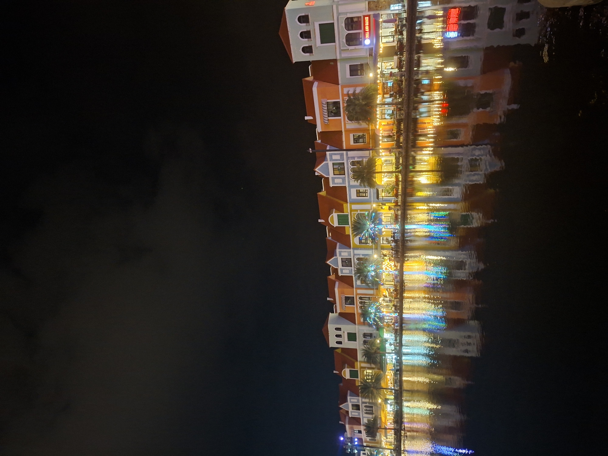 The image shows a row of colorful buildings reflected in calm water at night. The buildings are multi-storied with varying architectural styles and are illuminated by artificial lights. Each building is painted in distinct, vibrant colors such as orange, blue, and white, with red tiled roofs. Many windows are visible, some with lights on inside, indicating occupancy or activity. Palm trees, some adorned with colorful string lights, are interspersed among the buildings. The water surface is smooth, creating clear reflections of the buildings and their lights, which appear as elongated streaks of color. The scene is dark, with the exception of the artificial illumination from the buildings and their reflections. The sky is dark and appears cloudy, suggesting a night time setting. The overall atmosphere is festive and tranquil, evoking a sense of a vibrant waterfront area at night. One sign is visible on one of the buildings, partially obscured by reflection, reading "CHILI AND DANCE". Based on the location context of Phú Quốc, Vietnam, this scene likely depicts a waterfront promenade or entertainment district.