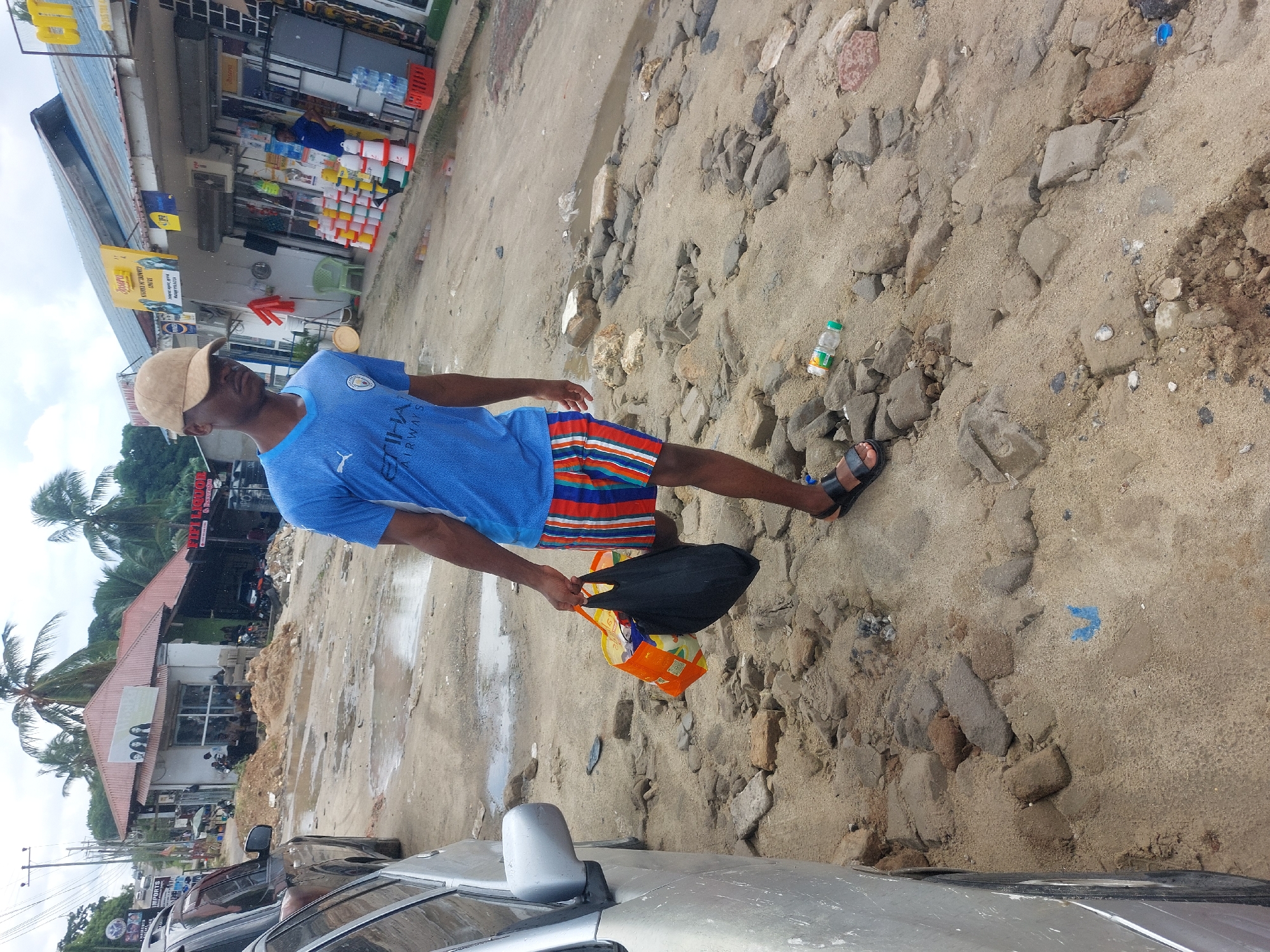 A man wearing a blue t-shirt, striped shorts, and sandals stands on a sandy, rocky surface near a street in Dar es-Salaam, Tanzania. He is holding shopping bags. The background shows buildings, palm trees, parked cars, and commercial establishments, one of which appears to be a retail store. The environment suggests a coastal or urban area undergoing construction or renovation.