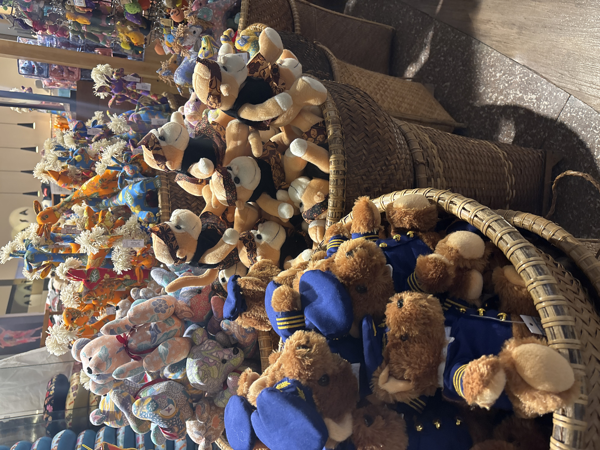 A high-angle indoor shot depicts an assortment of stuffed animals filling several wicker baskets. The subjects range from giraffes and bears to dogs, with varied patterns and colors covering their fabric exteriors. One basket is filled with brown bears dressed in blue uniforms. The baskets are densely packed, creating a colorful and textural scene. A partial reflection of merchandise and overhead lighting is visible on the left side of the frame, hinting at a store setting. The ground appears to be tiled in a neutral tone.
Based on the merchandise often sold there, it is possible that this scene is in Kuta, Indonesia.