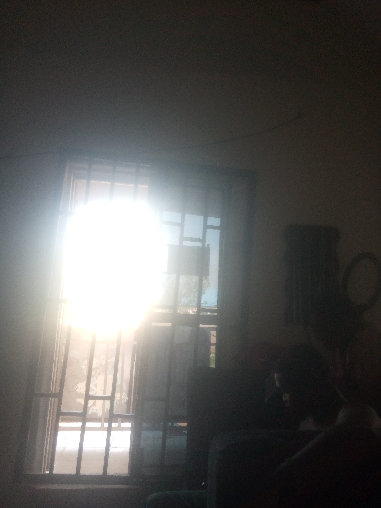 The image depicts an indoor scene, likely a residential interior, with a prominent window covered by a security grate. Intense sunlight floods through the window, creating a strong lens flare and washing out details of the exterior view. The interior is dimly lit, with the exception of the light from the window. In the foreground, partially obscured by shadows, are silhouettes of people and furniture. To the right, a circular object, possibly a mirror or a decorative item, is visible on the wall. The overall impression is of a space with limited natural light, where the sun's brightness is a dominant visual element. The location is identified as Jalingo, Nigeria.