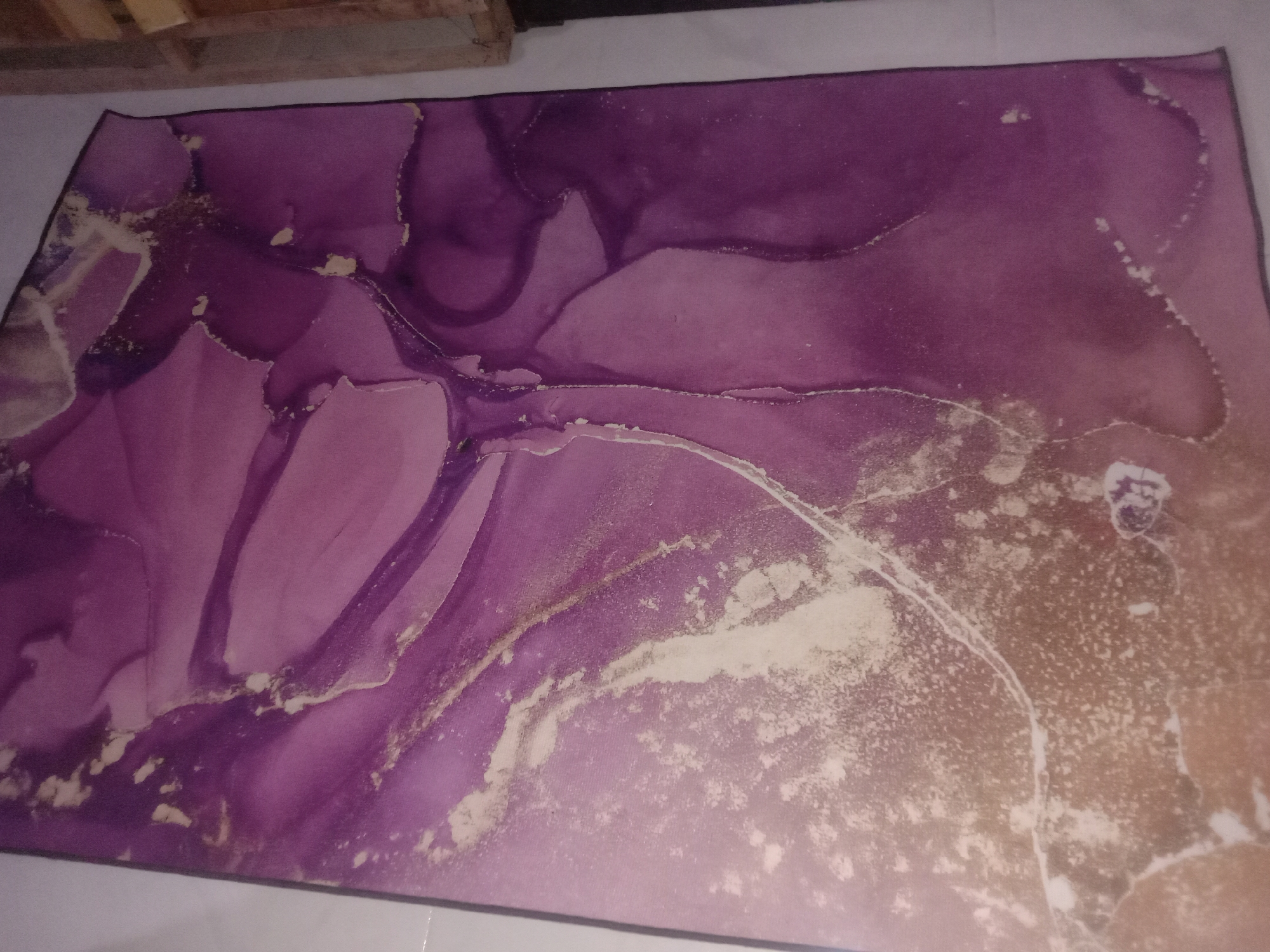 The image shows a rectangular mat with a marbled pattern in shades of purple, white, and gold. The pattern appears abstract, resembling fluid art. The mat is placed on a white tile floor. A wooden structure is partially visible in the upper left corner of the frame. No people are visible. The location, Lhokjok, Indonesia, is not discernible from the image.