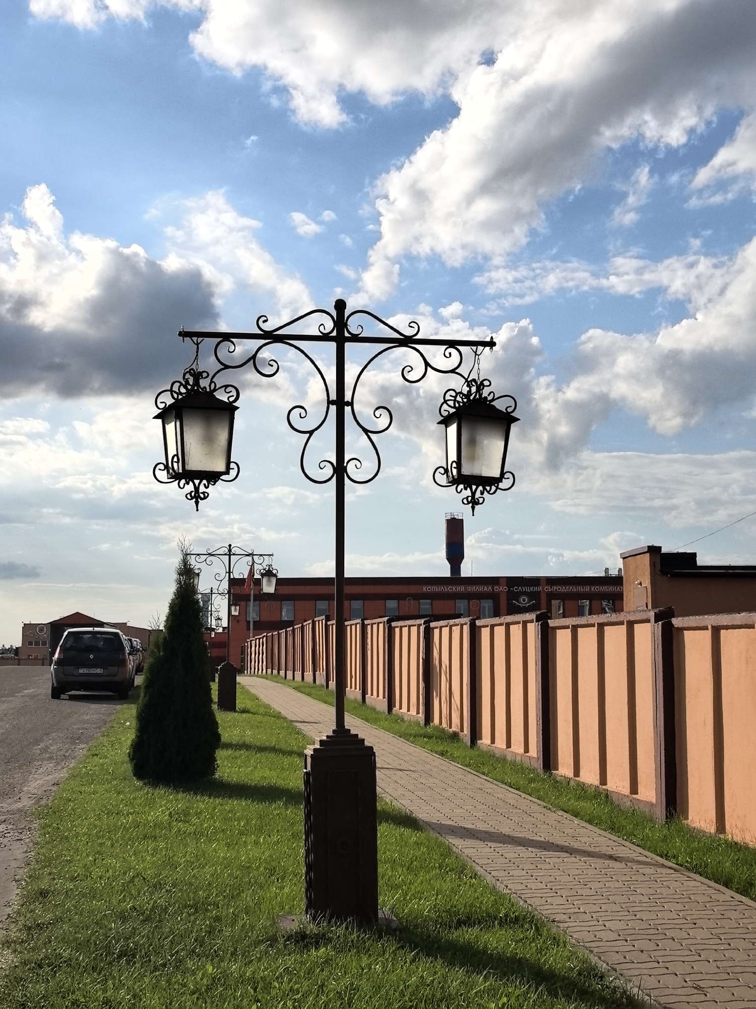 In the city of Unknown, Unknown, a street scene is captured under a sky filled with white, puffy clouds. The foreground features a black, ornate lamppost with two decorative lanterns hanging from curved arms. To the left of the lamppost, a patch of vibrant green grass slopes gently, with a dark green, conical evergreen tree positioned near its center. Beyond the grass, a paved walkway runs parallel to a tall, orange fence with dark vertical posts. Several cars are parked on the left side of the frame, with the closest being a dark-colored hatchback. In the background, a long, orange building with dark-trimmed windows is visible, and further beyond that, a tall, cylindrical structure with red and blue bands stands against the sky. The scene appears to be an outdoor public area, possibly a park or walkway adjacent to a commercial or industrial facility.