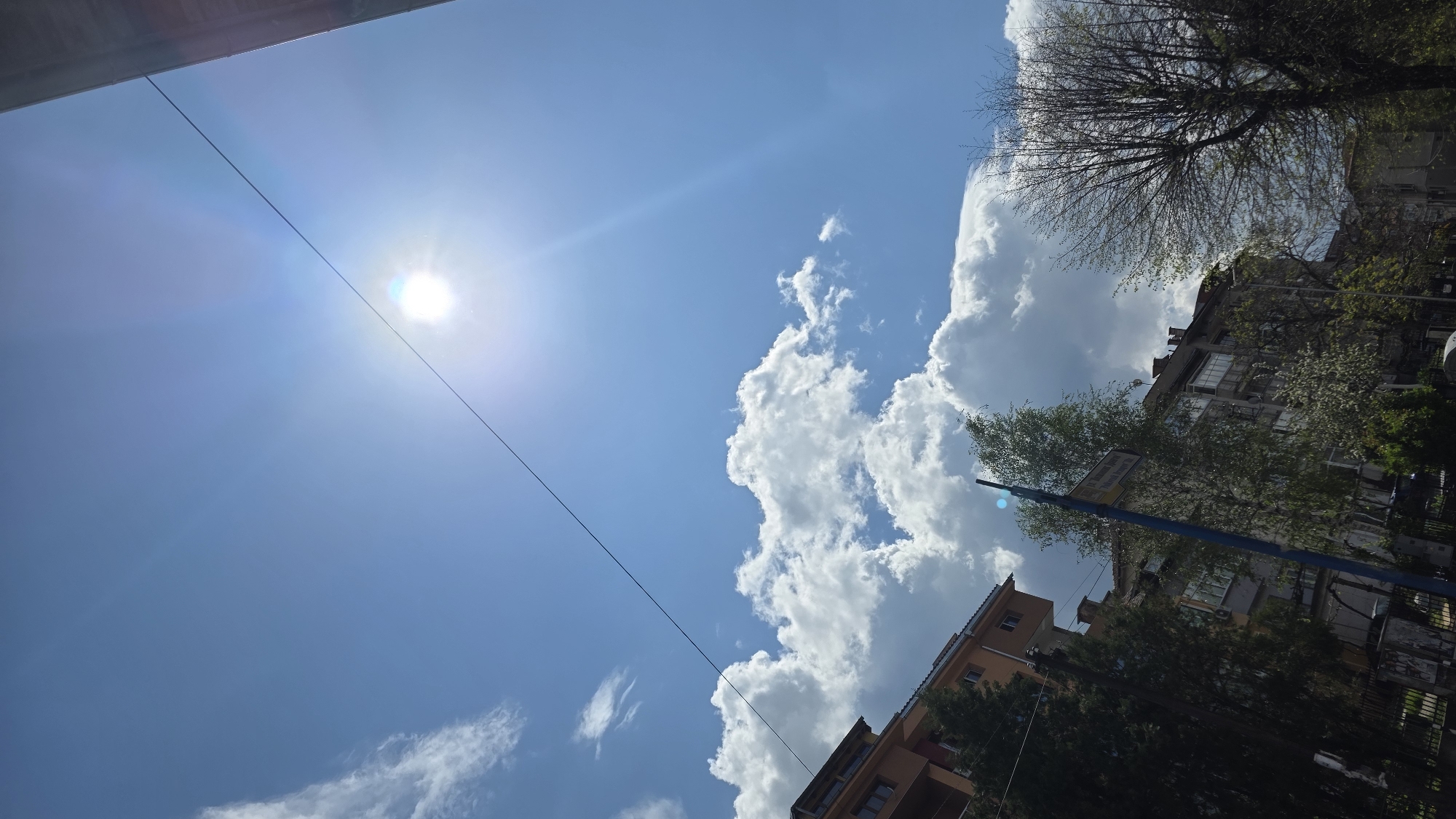 The image is a tilted, upward-looking shot of a partly cloudy sky in what appears to be an urban environment. The sun is visible in the upper left of the frame, partially obscured by a powerline running diagonally across the image. In the right side of the image, a building with orange walls is observed, and the outlines of trees are noticeable against the background of white, cumulus clouds.
Given the title details provided, the location is Blagoevgrad, Bulgaria. However, without further information, precise identification within the city is not possible.