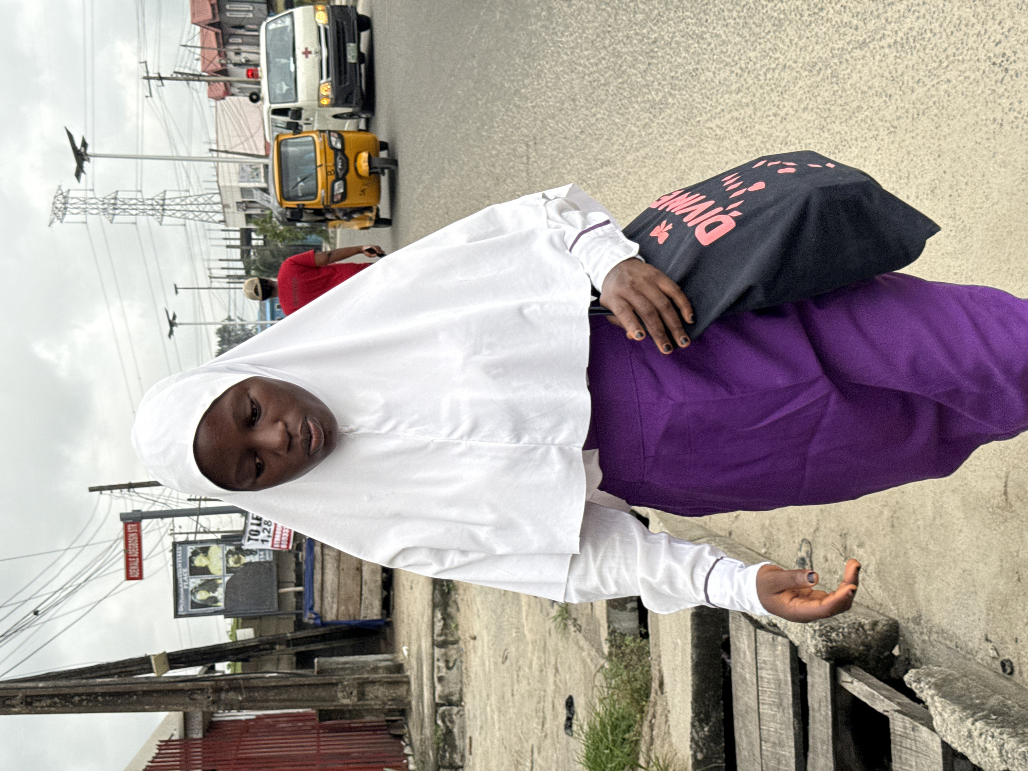 A young person, likely female, stands on a paved surface beside a street in Ado, Nigeria, under an overcast sky. She has dark skin and wears a white hijab over a long-sleeved white top, paired with a vibrant purple skirt or dress. Her expression is neutral as she looks slightly downwards. In her left hand, she carries a black bag adorned with the pink text "DIVINE" and a pink floral graphic. Her right hand is extended forward, palm slightly open, revealing fingernails painted with a dark polish.
The background depicts a busy urban street scene. On the paved road, a white ambulance with a visible red cross symbol is seen, followed closely by a yellow Keke Napep (auto-rickshaw). Numerous utility poles crisscross the sky with a network of wires, leading to a prominent, large metal electricity pylon in the distance. Along the roadside, various buildings with red-tiled roofs are visible.
Visible text on a street sign clearly reads "ADENIYI ADEGBOSUN STR." Another billboard or sign displays partial text such as "TO LET 1,2,8..." alongside images of people. A person wearing a red shirt and a light-colored cap is partially visible in the mid-ground, walking towards the road. The immediate foreground features concrete ledges and some weathered wooden structures, with sparse green vegetation growing nearby. The scene captures a moment of daily life in a bustling Nigerian street.
