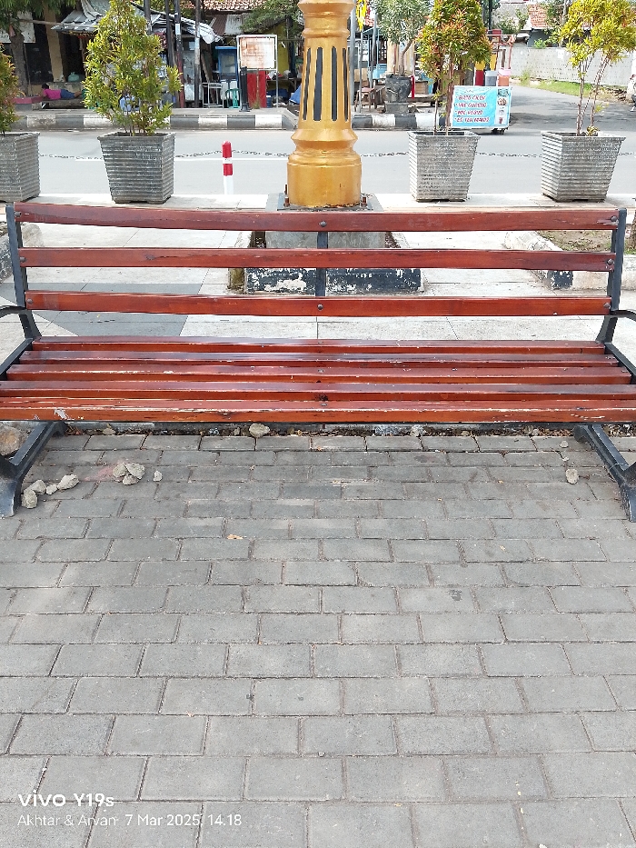 The image shows a wooden bench with black metal supports. The bench is empty and situated on a gray brick sidewalk in front of a gold-painted pole. There are two potted plants, one on each side of the bench, in front of the pole. The sidewalk is made of rectangular bricks with a slight gray patina. The bench has a wooden back and a seat made of slats. There are a few pebbles scattered around the base of the bench. The location is in Indramayu, Indonesia, at latitude -6.326, longitude 108.323. The image is taken from a low angle looking down at the bench. The bench is located in a public space. The bench is clean and well-maintained.