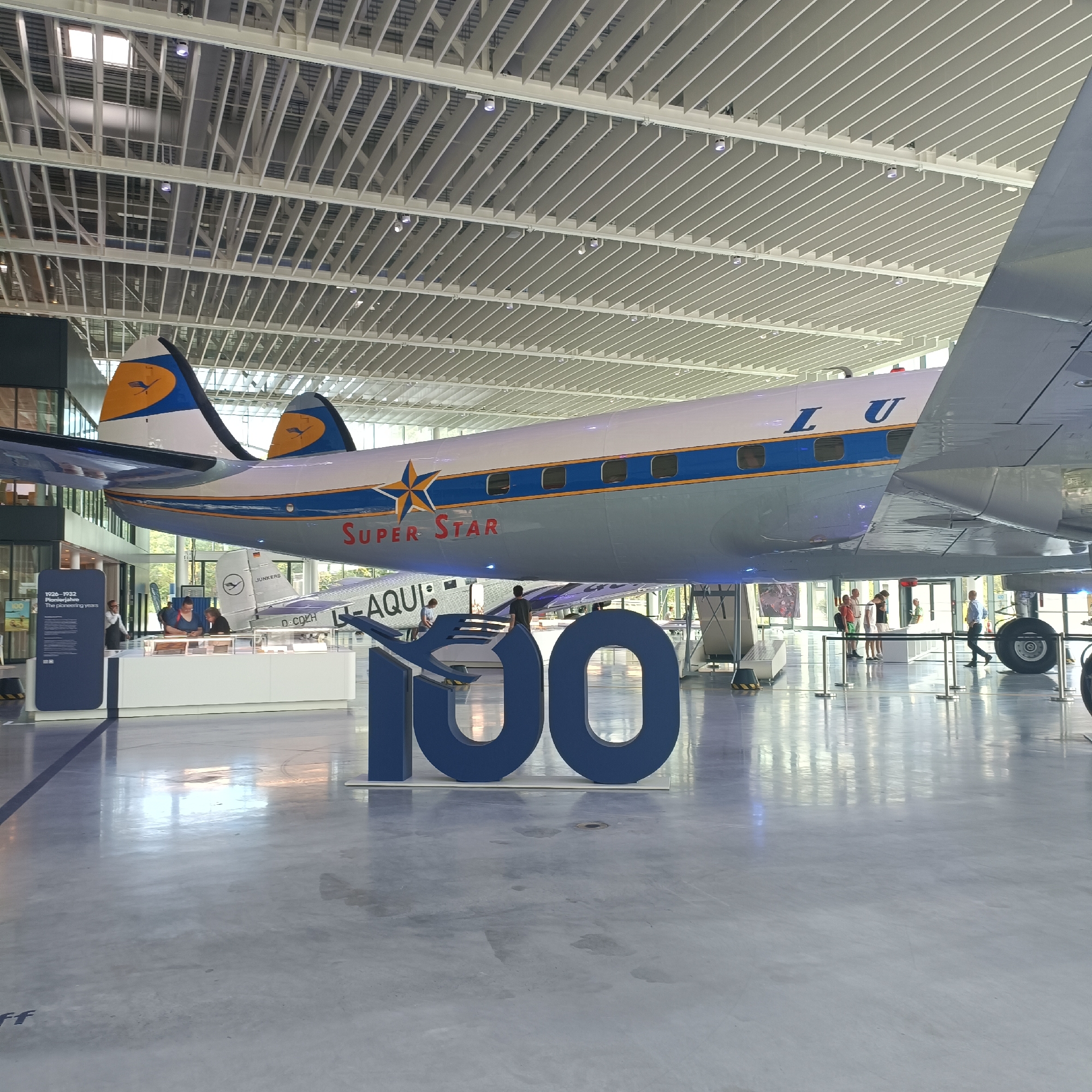 The image shows the interior of a museum, possibly an aviation museum. In the foreground, a large blue number "100" is displayed on the polished floor. Behind the number, an airplane is suspended from the ceiling. The airplane is white with blue and yellow accents, and the words "Super Star" are visible on its side. Another airplane, a silver propeller plane with the registration D-CDLH, is visible beneath the suspended aircraft. Several people are scattered throughout the museum, some looking at displays and others observing the aircraft. The ceiling of the museum is a complex structure of white beams and panels. The environment is well-lit with artificial lighting. It is likely the Lufthansa Aviation Center or a similar aviation exhibition space in Frankfurt, Germany, given the presence of Lufthansa branding on the aircraft.