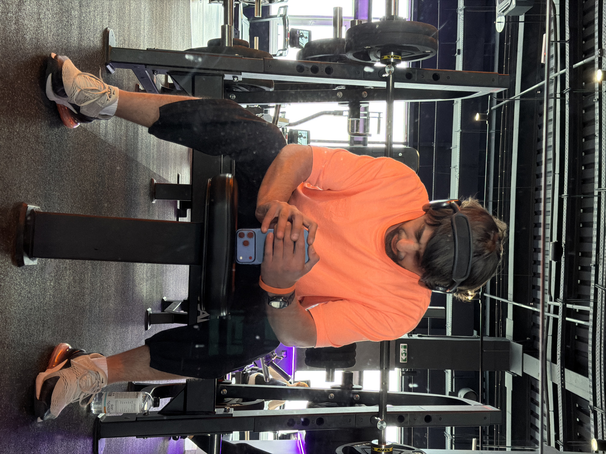 A man is seated on a leg extension machine in a gym. He is wearing an orange t-shirt, black shorts, white athletic shoes, and headphones. He is holding a blue smartphone in his hands. A water bottle sits on the machine near his feet. The gym has a black and gray color scheme, with exposed metal beams and weights visible. The location cannot be determined to be in Krasnoyarsk, Russia, without further information.