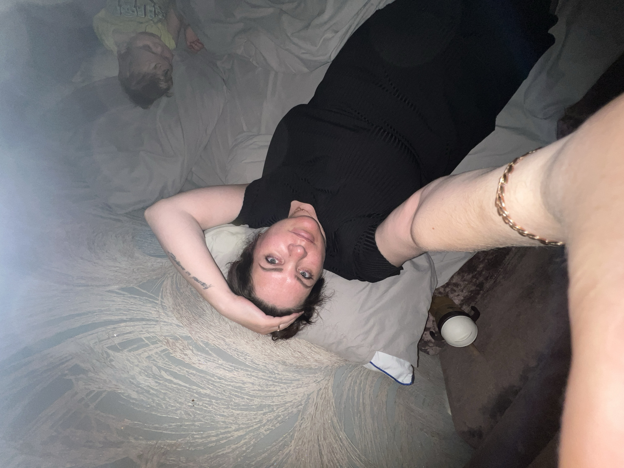 A woman in a black dress lies on a bed with a floral patterned bedspread. A young child lies near the top left corner of the image, seemingly asleep. The woman has her hand on her head and faces the camera, taking a selfie. A water bottle is on the bed near her torso. An arm with a gold bracelet is visible in the foreground. The location is not discernible from the image.