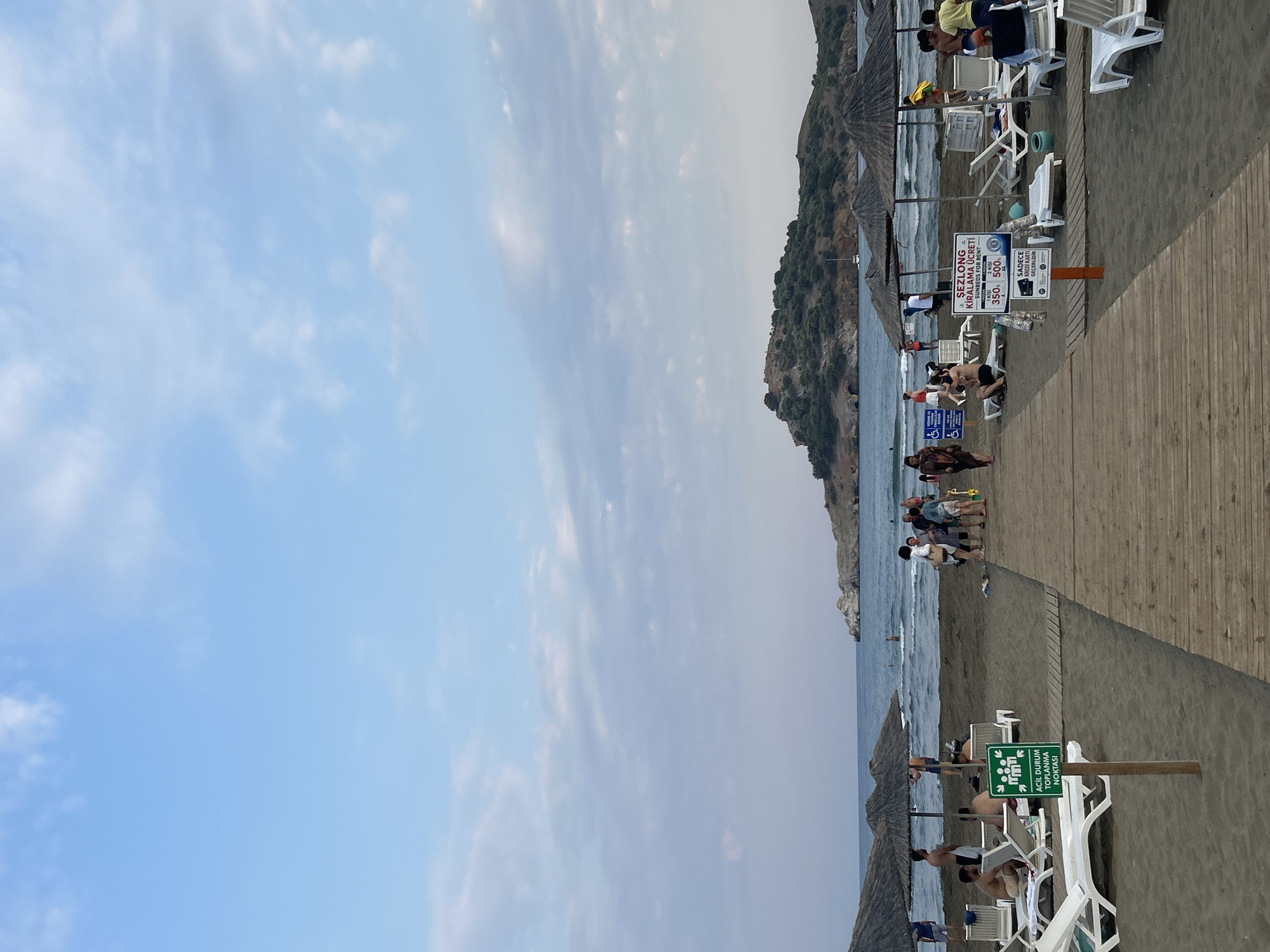 The image depicts a beach scene in Ortaca, Turkey. The foreground shows a sandy beach with a wooden walkway leading towards the sea. Numerous beach chairs and umbrellas are set up on the sand. A few people are visible lounging on the chairs, walking on the beach, or in the water. Signs are present, one with an "acil durum toplanma noktasi" (emergency assembly point) symbol and another indicating "sezlong kiralama ucreti" (sunbed rental fee) with prices listed in Turkish Lira. In the mid-ground, the sea is visible with gentle waves and a few people swimming. In the background, a rocky, tree-covered island is situated in the sea. The sky is a pale blue with scattered white clouds, suggesting it is either early morning or late afternoon. The overall atmosphere is relaxed and typical of a beach resort.