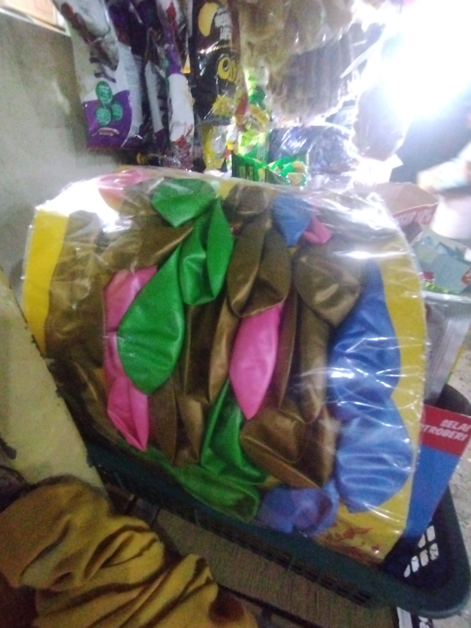 The image depicts a package of uninflated balloons of various colors (green, pink, brown, and blue) inside a clear plastic bag with a yellow border, located in what appears to be a small shop or stall. Other items such as snacks and a "selai stroberi" (strawberry jam in Indonesian) product are visible in the background, suggesting a retail environment. The image was likely taken in a city, given the presence of retail goods. If this is in the city of Lhokseumawe, Indonesia, it would be reasonable to assume the stall is selling items that would be commonly found in such an environment.