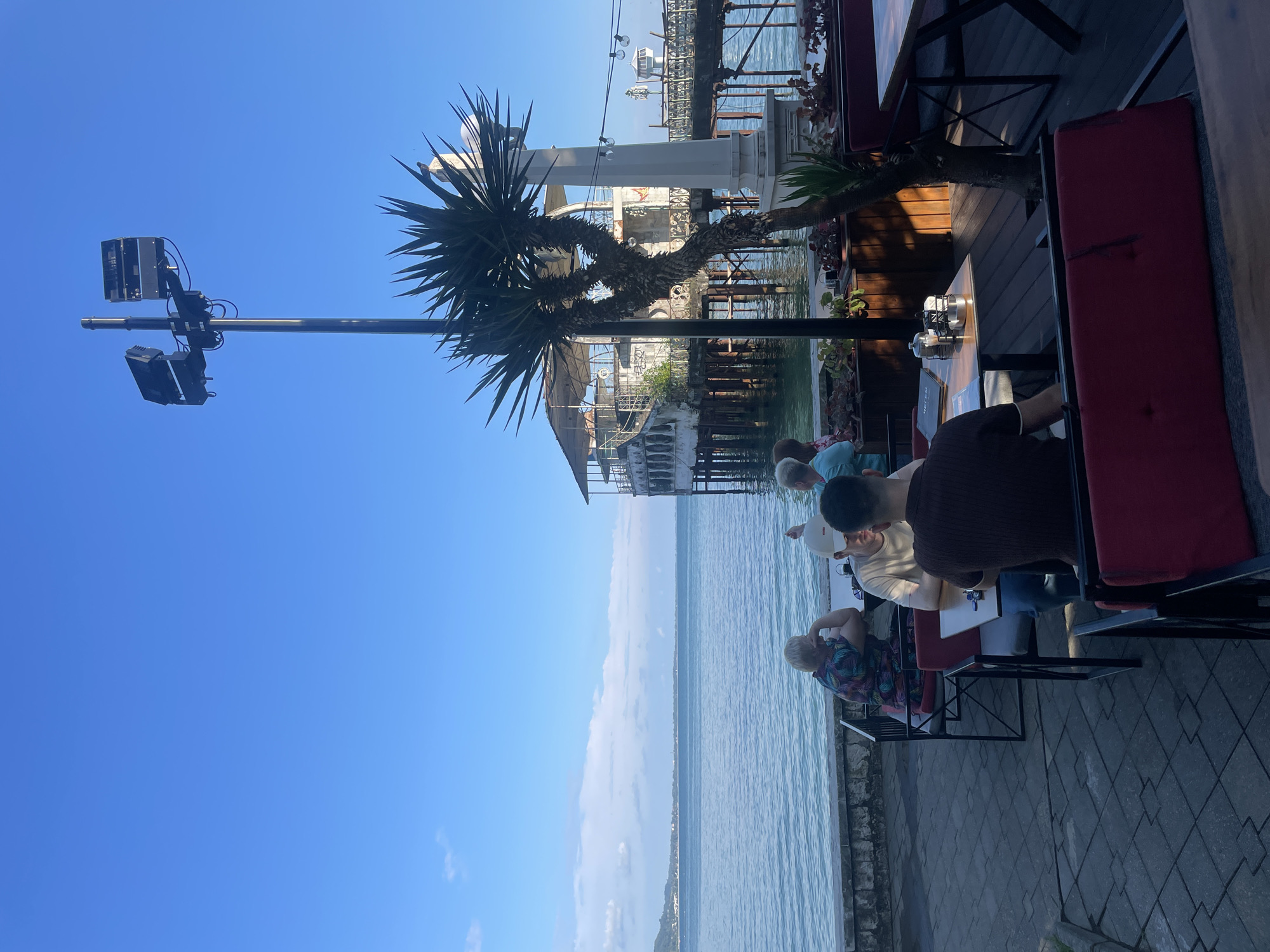 The image depicts an outdoor seating area of a restaurant or cafe situated along a waterfront in Sokhumi, Abkhazia. In the foreground, several individuals are seated at tables. One person, wearing a colorful patterned shirt, is seen with their head resting on their hand, looking out towards the water. Adjacent to them, two men are seated at a table, one wearing a light-colored t-shirt and a cap, and the other facing away from the camera, wearing a dark-colored polo shirt. Red upholstered benches are visible in the immediate foreground.
The seating area overlooks a body of water, likely the Black Sea, with a coastline and distant landforms visible on the horizon. A stone wall separates the paved patio area from the water's edge. In the mid-ground, a distinctive structure, an old, dilapidated building on stilts over the water, possibly a former restaurant or attraction, is prominent. A palm-like tree stands to the right of this structure. A tall pole with multiple lights or speakers is also visible, dominating the skyline. The sky is a clear, bright blue with a few wispy clouds. The overall scene suggests a tranquil, late afternoon or early evening setting.