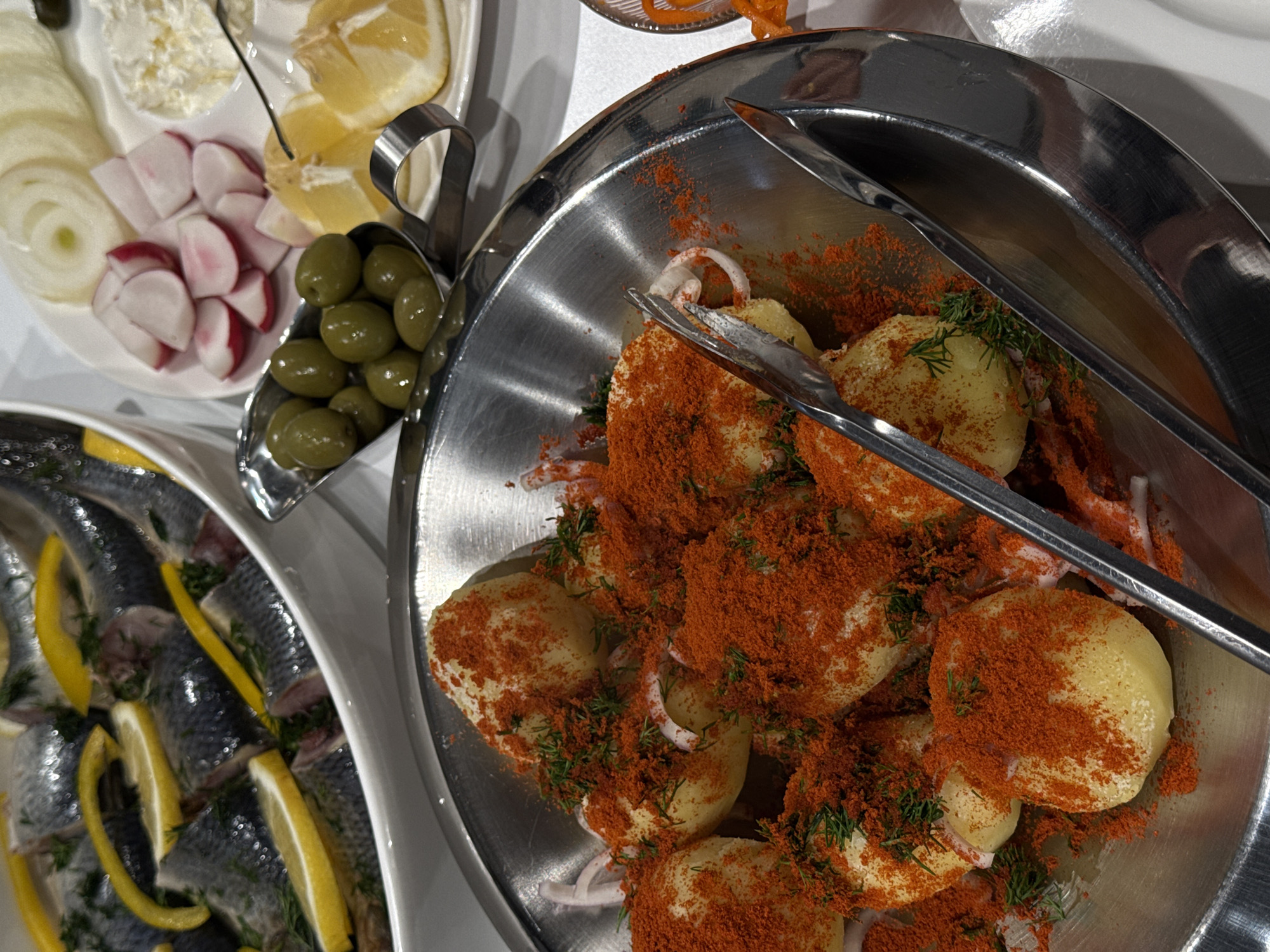 The image shows a spread of food items. A silver bowl contains potatoes sprinkled with red powder, possibly paprika. There are sliced onions and herbs mixed in. Silver tongs rest in the bowl. To the left, a white plate holds sliced onions, radishes, a creamy white substance, lemon wedges, and green olives in a small silver dish. A separate white platter contains herring with lemon slices and dill. The setting appears to be an indoor buffet or dining establishment. The city and country, Yerevan, Armenia, are inferred.