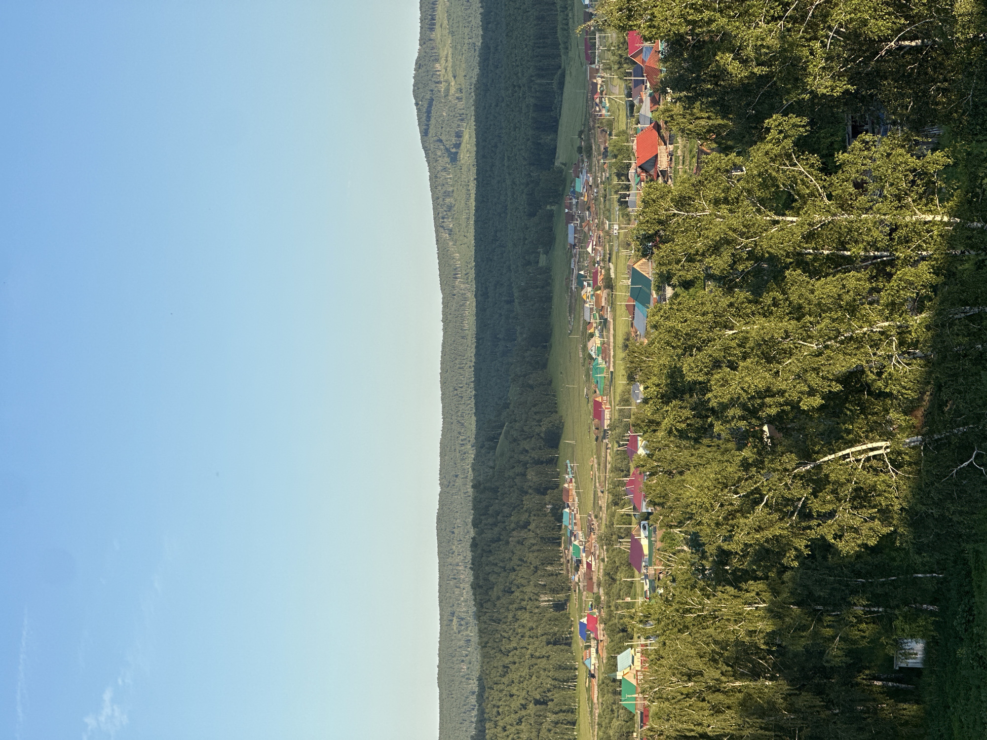 This is an aerial photograph of a rural village nestled in a valley surrounded by rolling, tree-covered hills. The sky is clear and blue with a gradient of lighter blue near the horizon. The foreground is dominated by lush green trees, predominantly birch trees with their distinctive white bark, casting shadows across the scene.
In the middle ground, the village unfolds, characterized by numerous houses with brightly colored roofs in shades of red, green, blue, and orange. The houses appear to be single-story structures with visible chimneys and fences. Some of the houses are clustered together, while others are more spread out. Roads or paths wind through the village.
The background features layers of dense green forests covering the hills, with a hazy, lighter green layer indicating more distant hills. The overall impression is one of a peaceful, pastoral landscape bathed in the warm light of either early morning or late afternoon sun. There are no people visible in the frame. The environment appears to be natural and somewhat undeveloped, typical of a rural or semi-rural setting.