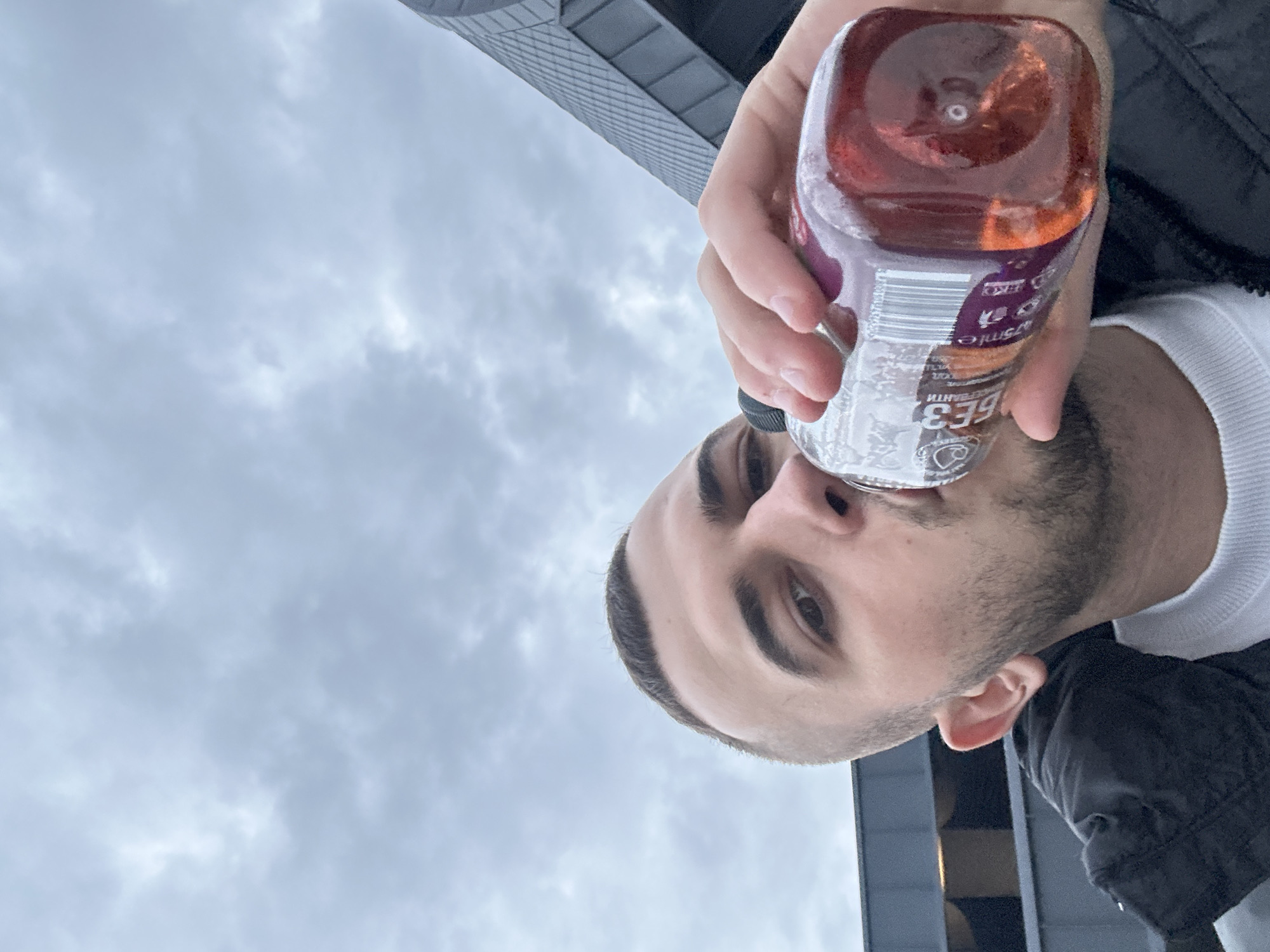 The image is a low-angle, upside-down shot featuring a young man drinking from a bottle, set against a cloudy sky and a modern building. The man, likely in his 20s or 30s, has short dark hair, distinct eyebrows, and a short beard. He is looking directly at the camera with a neutral expression. He wears a white crew-neck shirt beneath a dark jacket.
He holds a clear plastic bottle filled with a reddish-orange liquid. The bottle's label is partially visible, showing text in Cyrillic script, including "575ml", "БЕЗ ГМО" (meaning "GMO-free"), and "ПИТИЕ" (meaning "drink"). A barcode is also discernible on the label.
The background consists primarily of a cloudy, overcast sky, indicating a dull daytime. A modern building with a textured, grid-like facade is visible in the upper right portion of the background, suggesting an urban environment. This scene is situated in Plovdiv, Bulgaria.