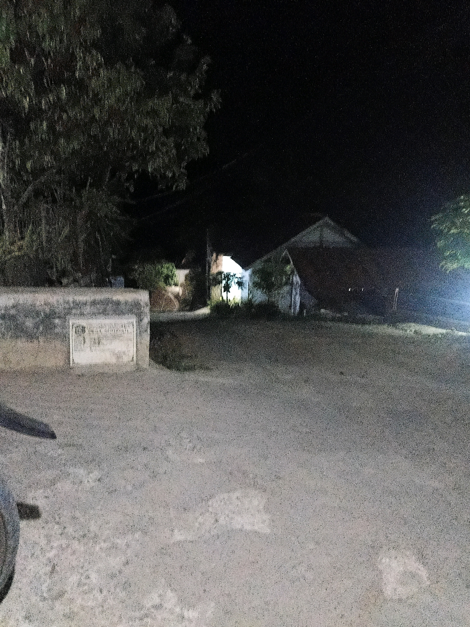 The image depicts a nighttime scene in the city of Sinduaji, Indonesia. The foreground is dominated by a dusty, grey road. To the left, a low, grey concrete wall is visible, with a plaque or sign embedded in its surface. This plaque appears to be made of a lighter colored material, possibly stone or metal, and contains some text, though it is largely illegible due to the low light and image quality. Behind the wall, a large tree with dark foliage is present. Further back, several buildings are dimly lit. One structure has a pitched roof and appears to be made of a light-colored material. Another building or structure to the right is partially illuminated by an external light source, casting a bright glow. The overall impression is of a quiet, rural or semi-rural area at night.