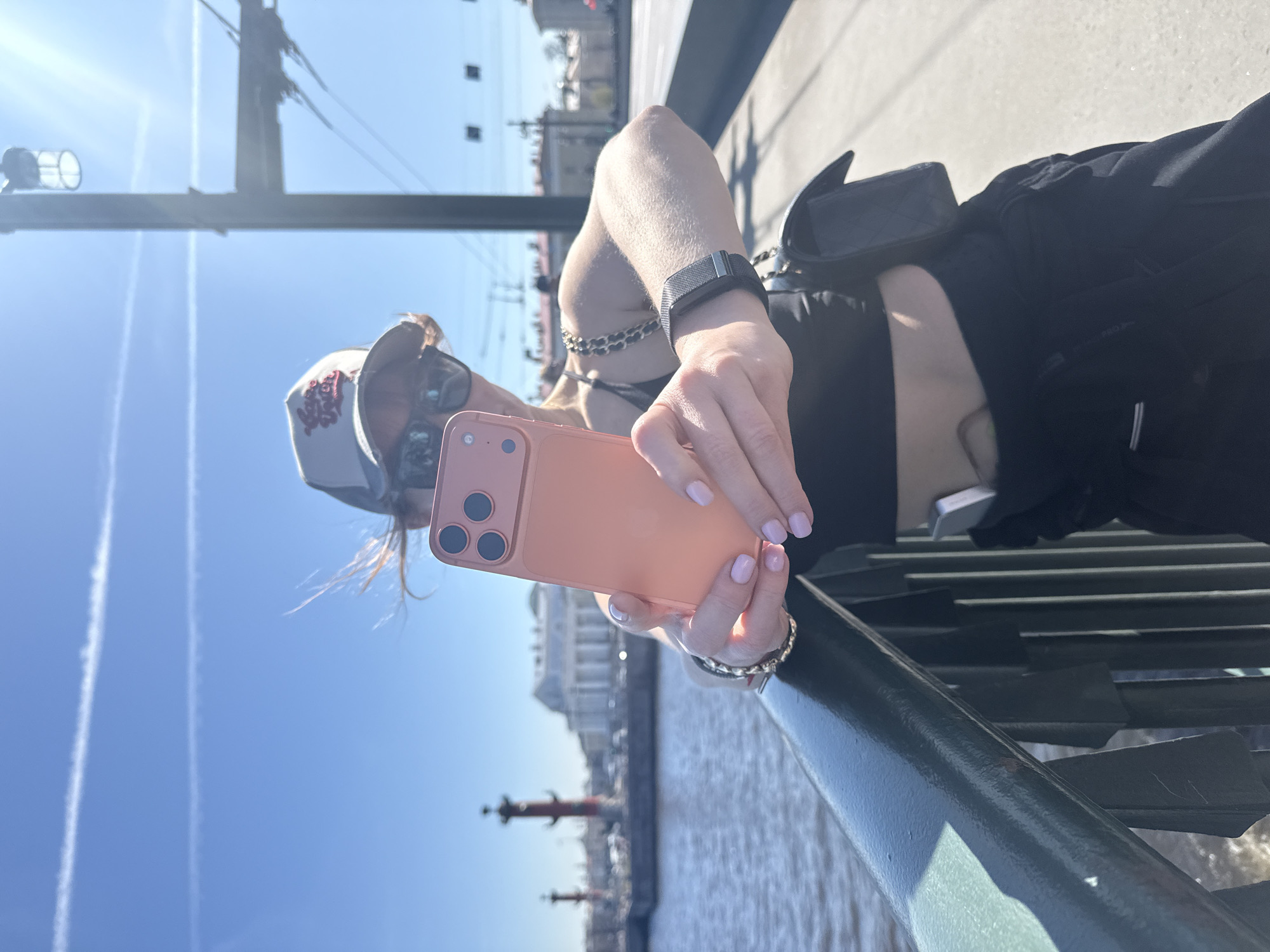 A woman holds an iPhone with a peach-colored case in her right hand. She is wearing a cap, sunglasses, a black top, and a black crossbody bag. A fitness tracker is visible on her left wrist. The background shows the Rostral Columns in Saint Petersburg, Russia, with a river and buildings. The sky is blue. The woman is standing on a bridge with a dark green railing.