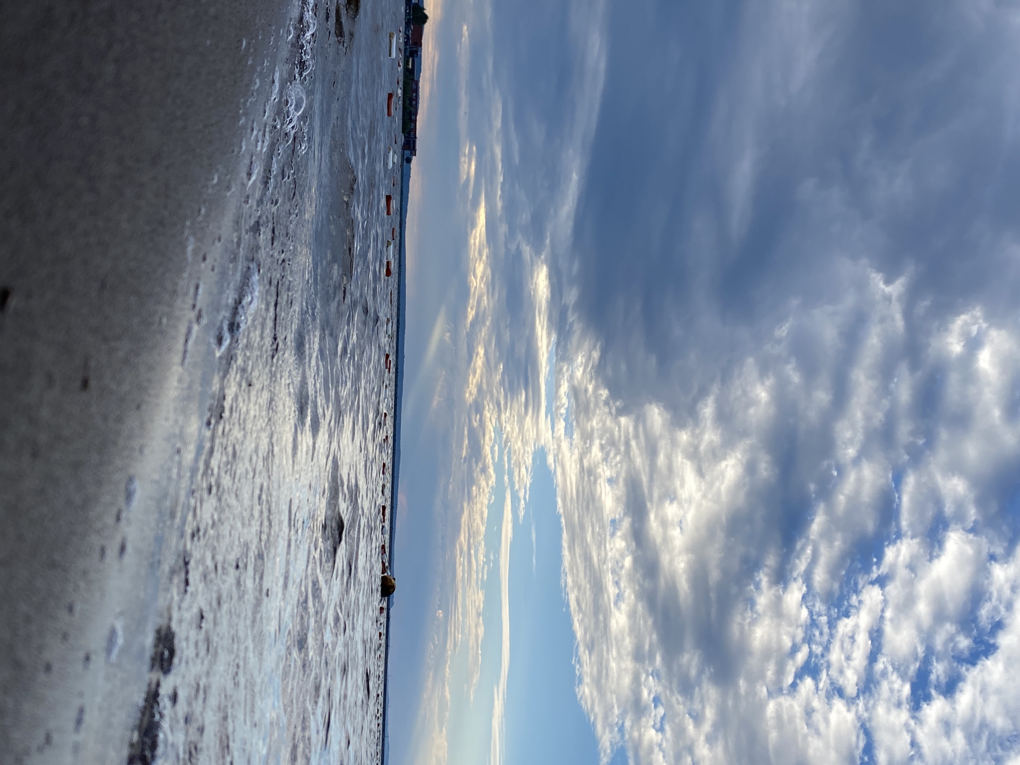 The image depicts a low-angle view of a body of water, likely a reservoir or large river, with a shoreline in the foreground. The water is choppy, with small waves and some foam and bubbles visible near the shore. In the distance, a line of buoys, some white and some orange, marks an area on the water. Beyond the buoys, a long, low-lying building or complex with a red roof is visible on the far shore. The sky is filled with a mix of dark, dramatic storm clouds and brighter, scattered clouds, with shafts of sunlight breaking through in places. The scene is consistent with the Kama River Reservoir, specifically in the vicinity of the city of Chaikovsky, Russia, known for its industrial and hydroelectric facilities along the river. No people are visible in the image.