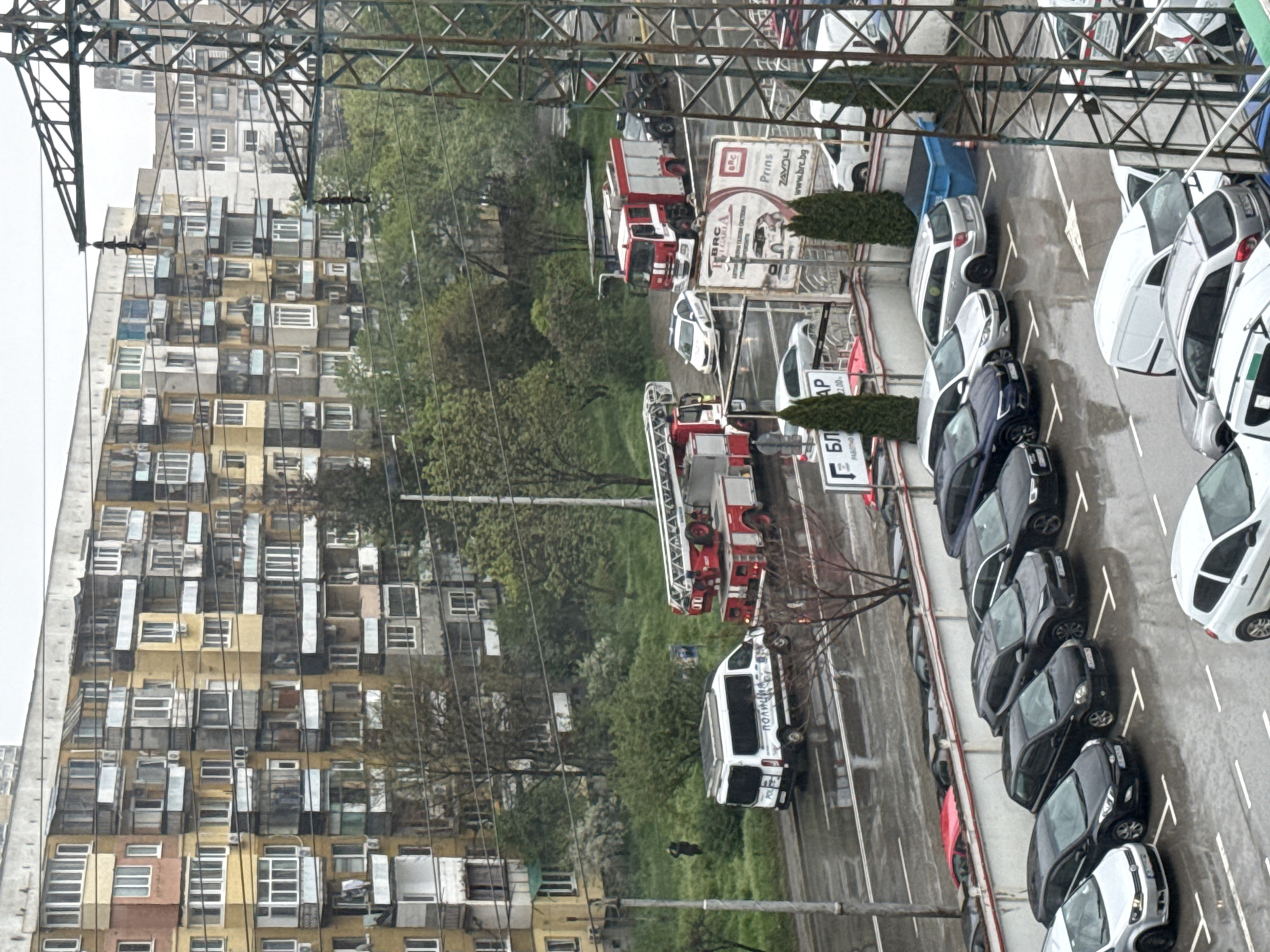 This elevated daytime image captures an urban scene in Sofia, Bulgaria, featuring residential buildings, a street, and a parking lot, under an overcast sky with wet ground.
On the left, several multi-story residential buildings with numerous windows and balconies exhibit varied architectural elements. Green trees and foliage are interspersed between these structures and the street.
The foreground and right side of the image are dominated by a street and a parking lot filled with numerous cars, predominantly dark or silver/white sedans and vans. Several emergency vehicles are prominent: two red and white fire trucks, one of which has its large ladder extended or deploying, and a white police van clearly marked with "ПОЛИЦИЯ" (Police). Another red fire truck is visible further down the street, near a blue refuse skip.
A complex network of metal trusses and overhead wires, likely part of an electricity pylon or utility infrastructure, spans across the upper portion of the frame.
Visible text includes:
- On a large billboard: "BRC", "Prins", "ZAVOLI", "www.brc.bg", and "АВТО ГАЗОВИ СИСТЕМИ" (Auto Gas Systems).
- On a partially visible street sign: "ПАРКИНГ" (Parking) and "22.00".
- Another sign shows "БЛ".
The scene suggests an ongoing emergency response or a training exercise involving fire and police services. While no specific human activity is clearly discernible, the presence of emergency vehicles implies personnel involvement.