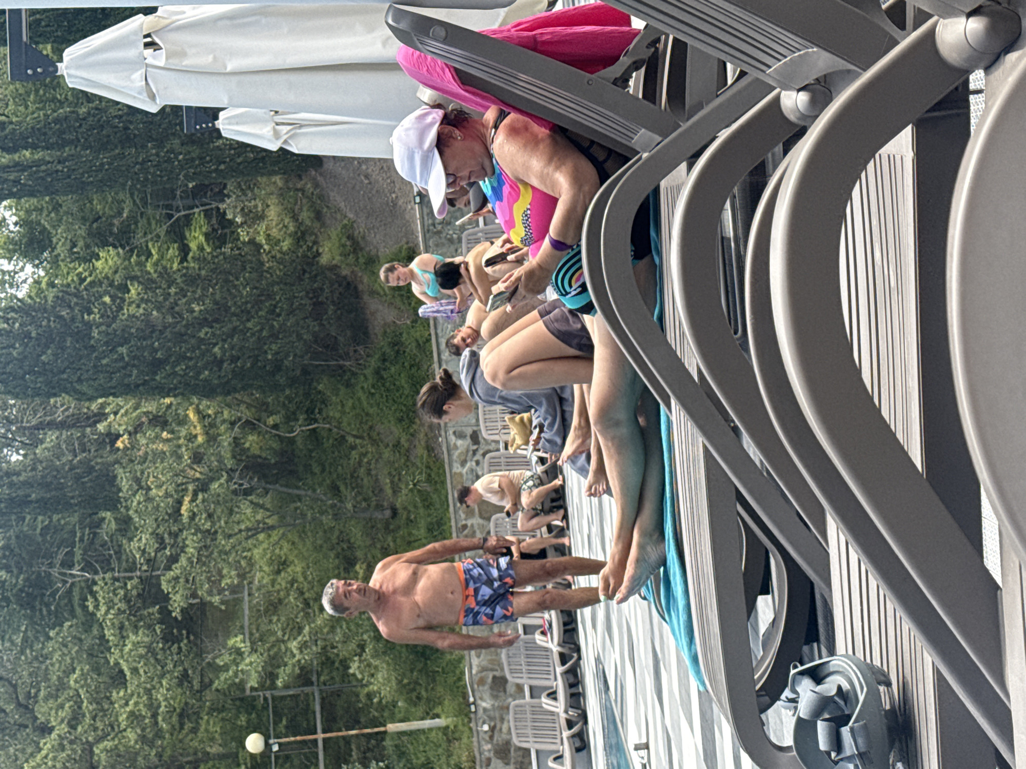 In Yalta, Ukraine, a scene unfolds by a swimming pool on a sunny day. An older man, shirtless and wearing patterned swim trunks, stands near the edge of the pool. Several people are relaxing on lounge chairs. In the foreground, a woman wearing a white cap and a colorful swimsuit is looking at her phone. Another person's legs are visible on a lounge chair draped with a blue towel. In the background, more lounge chairs and trees are visible. The environment appears to be a resort or hotel area.