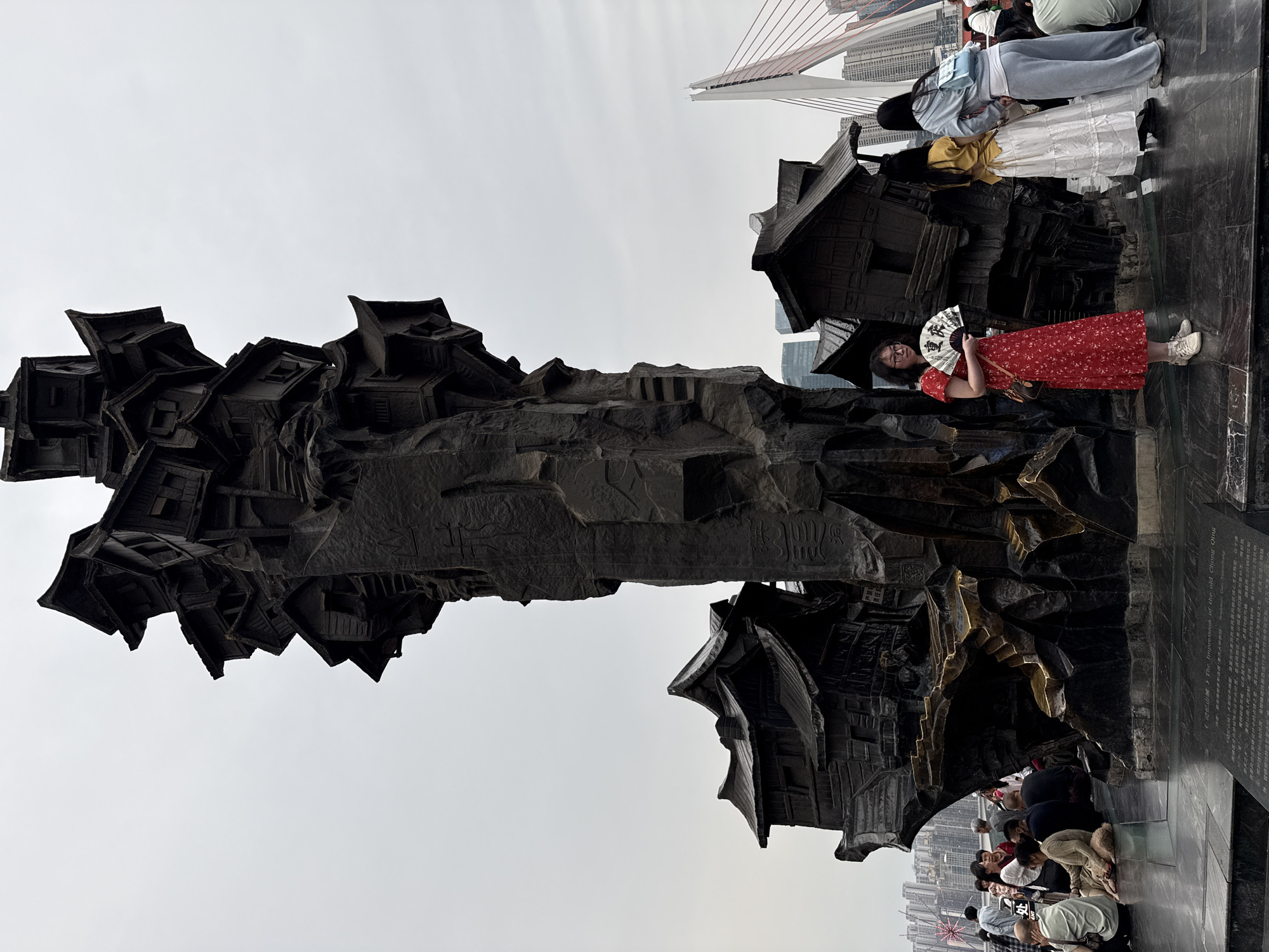 The image captures a low-angle, medium shot of a contemporary monument of stacked, angular buildings resembling traditional Chinese architecture. The monument is likely constructed from dark stone or metal, contrasting with the overcast sky. A woman in a red dress and white sneakers stands to the right, holding an opened decorative fan. Groups of people are clustered at the base of the monument, some kneeling or sitting and viewing the inscription on the plinth. A suspension bridge is visible in the background. Based on the inscription, the scene is of 'The Impression of the old Chong Qing', which is likely located in the city of Yuzhong District, China.