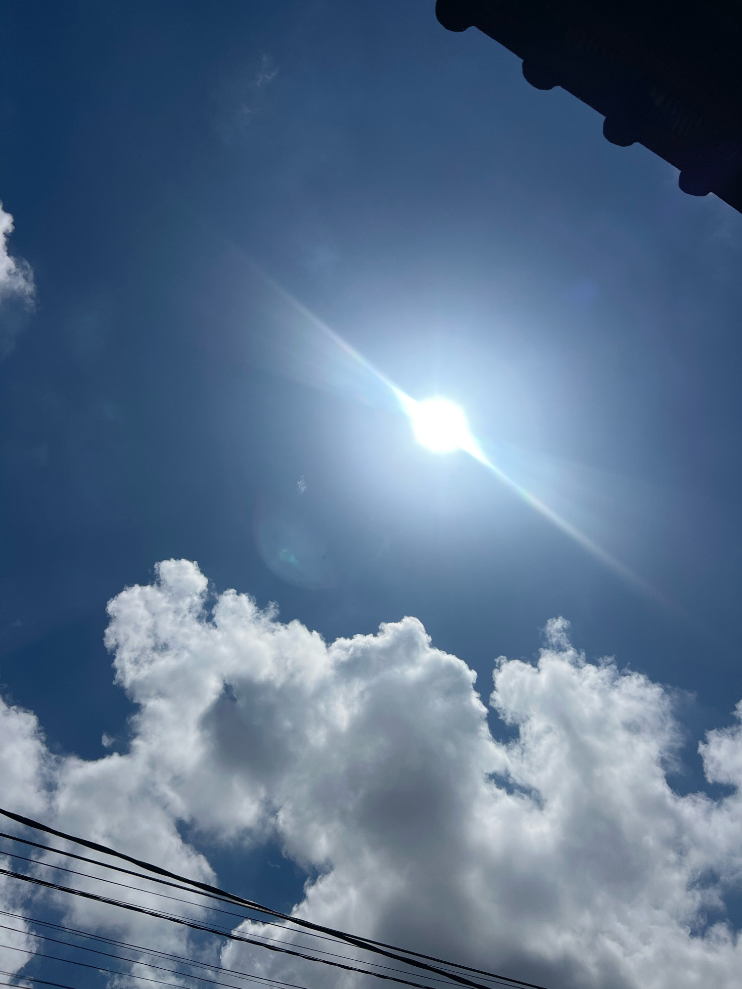 A low-angle shot captures a bright, sun-drenched blue sky in Denpasar, Indonesia. The sun is a brilliant white orb with a strong lens flare, casting a diagonal beam of light across the upper right quadrant of the frame. Fluffy white cumulus clouds are scattered across the sky, with a large cluster dominating the lower half of the image. A dark, corrugated roofline is visible in the upper right corner, silhouetted against the sky. Several black electrical wires run diagonally across the lower portion of the image, suggesting an urban or semi-urban environment. The overall impression is a clear, sunny day.