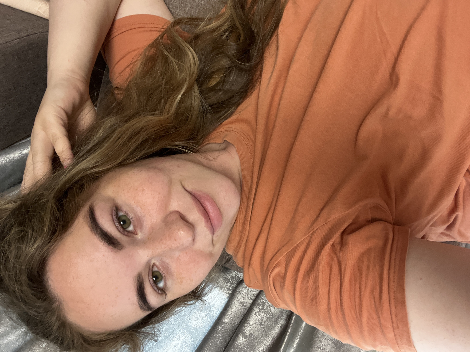 A female individual is depicted in a close-up, upward-angled portrait, situated in the city of Chaikovsky, Russia. She is lying on her side, looking directly at the camera with a slight, closed-lip smile. Her hair is medium-long, wavy, and multi-tonal light brown, draping over her right shoulder. Her face displays well-defined eyebrows, greenish-hazel eyes, and numerous freckles across her nose, cheeks, and forehead. Her lips are a natural light pink. She is wearing a plain, short-sleeved, crew-neck t-shirt in a solid orange color, with the fabric showing natural wrinkles. The left foreground is occupied by her arm and the orange t-shirt. The background, behind her head, consists of a textured, somewhat reflective fabric in shades of grey, silver, and light blue, indicative of an indoor environment, likely curtains or upholstery. Her right arm supports her head, and her left hand is partially visible near her hair on the right side of the frame.