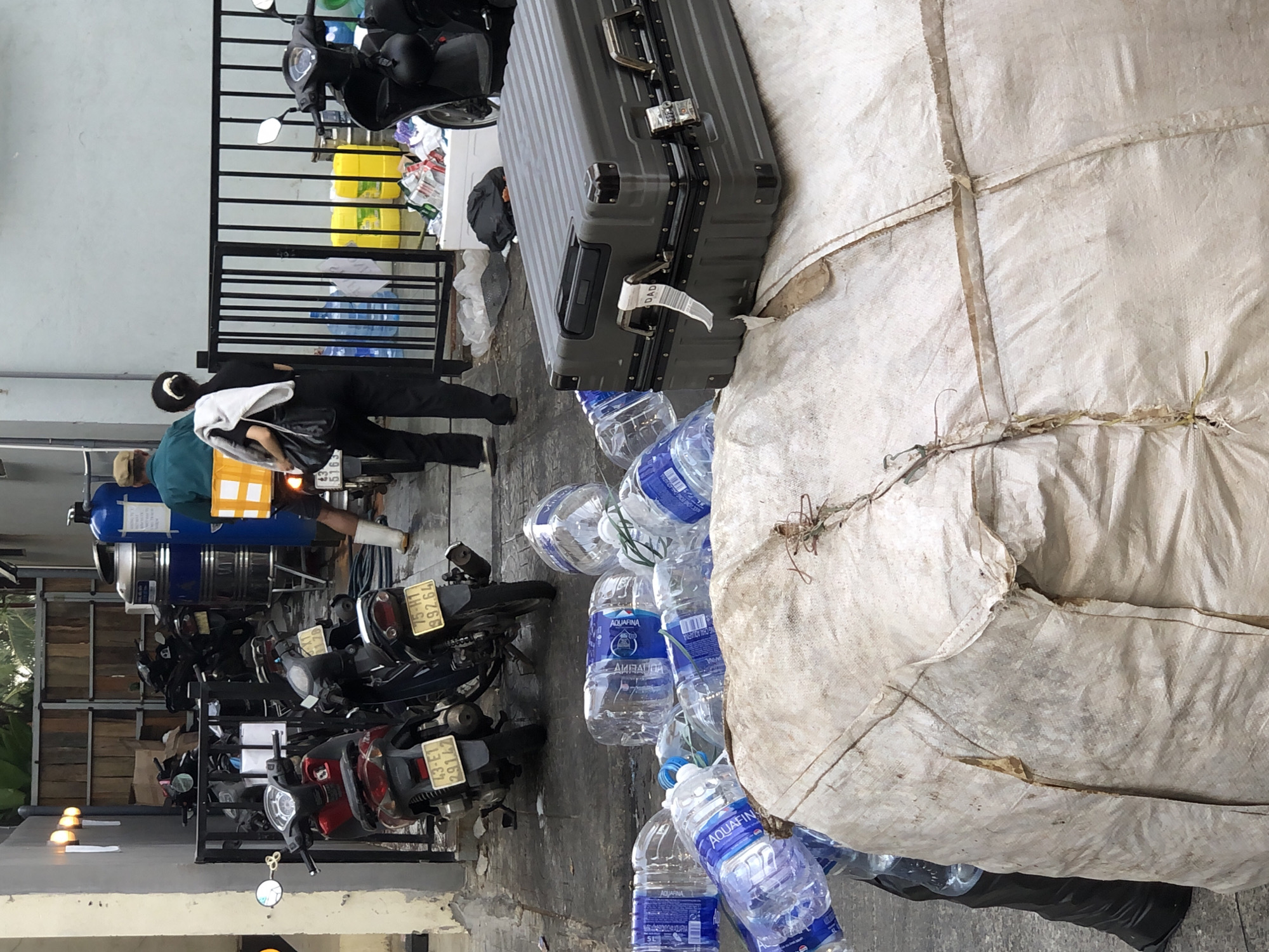 The image depicts an outdoor scene in what appears to be an alley or a service area in Đà Nẵng, Vietnam. In the foreground, there is a large, roughly woven sack, possibly burlap, partially obscuring a grey, hard-shelled suitcase with metal latches and a TSA lock. Several plastic water bottles, some labeled "AQUAFINA," are scattered around the sack and the suitcase. Behind these items, a black garbage bag is visible. Further back, a black metal fence separates the area from a walkway or street. Beyond the fence, a collection of motorbikes is parked. To the right of the motorbikes, a large blue water tank and some yellow containers are present. Two individuals are partially visible: one wearing a green jacket and a tan hat, standing near the water tank, and another person in dark clothing carrying a bag over their shoulder. The overall impression is one of a storage or collection point for various items, including water bottles and potentially recyclables. The environment appears to be utilitarian and possibly a loading or unloading zone.