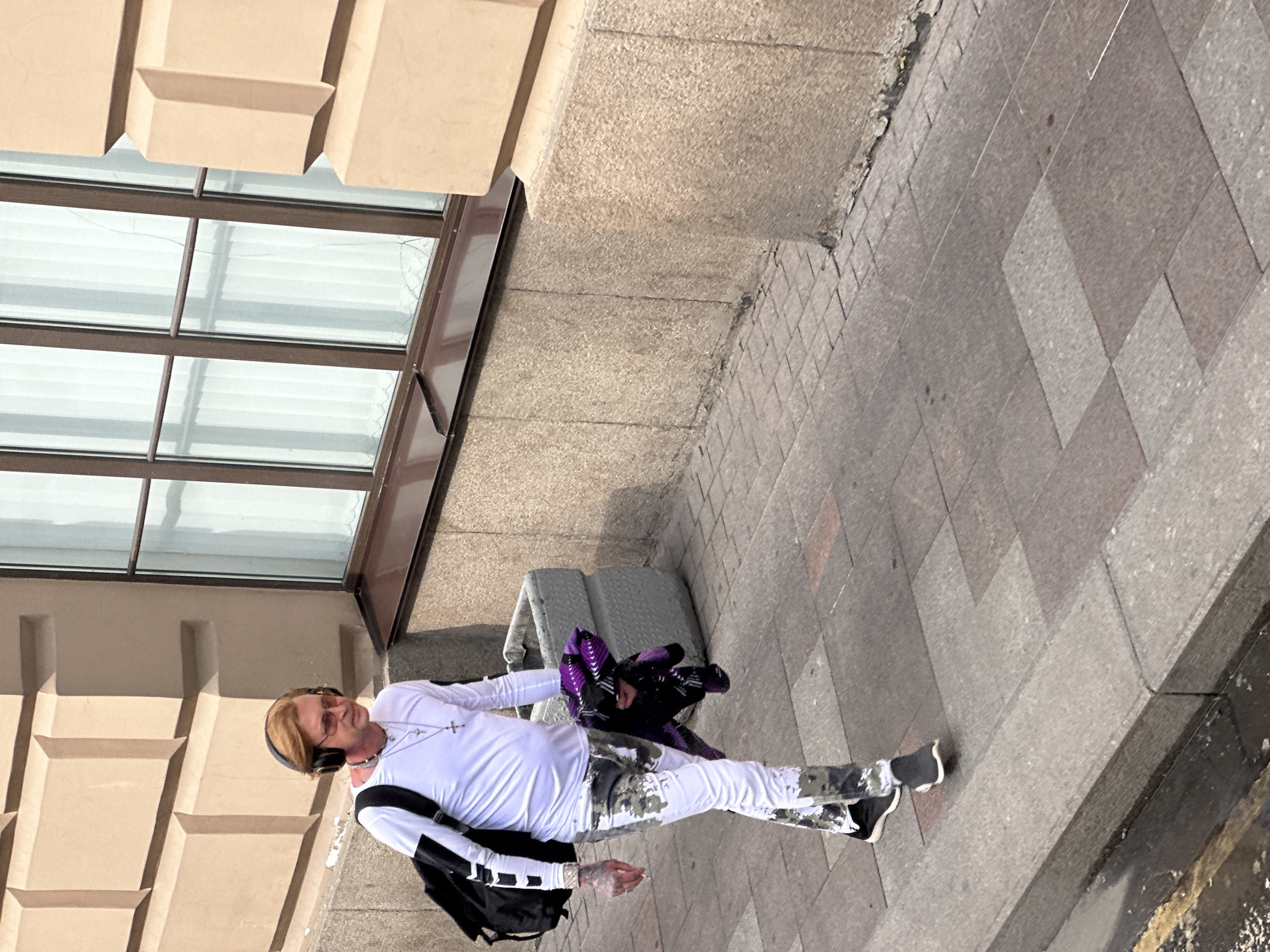 A full-body shot shows a man walking on a sidewalk in what appears to be Saint Petersburg, Russia. He is wearing headphones, a white long-sleeve shirt, camouflage pants, and black and white sneakers. He carries a black backpack on his left shoulder. In his left hand, he holds what appears to be a purple garment. He walks alongside a building with large windows. Next to the building is a gray object that may be a bench or an electrical box. The sidewalk is paved with various sizes of square and rectangular gray tiles. The perspective is slightly tilted.