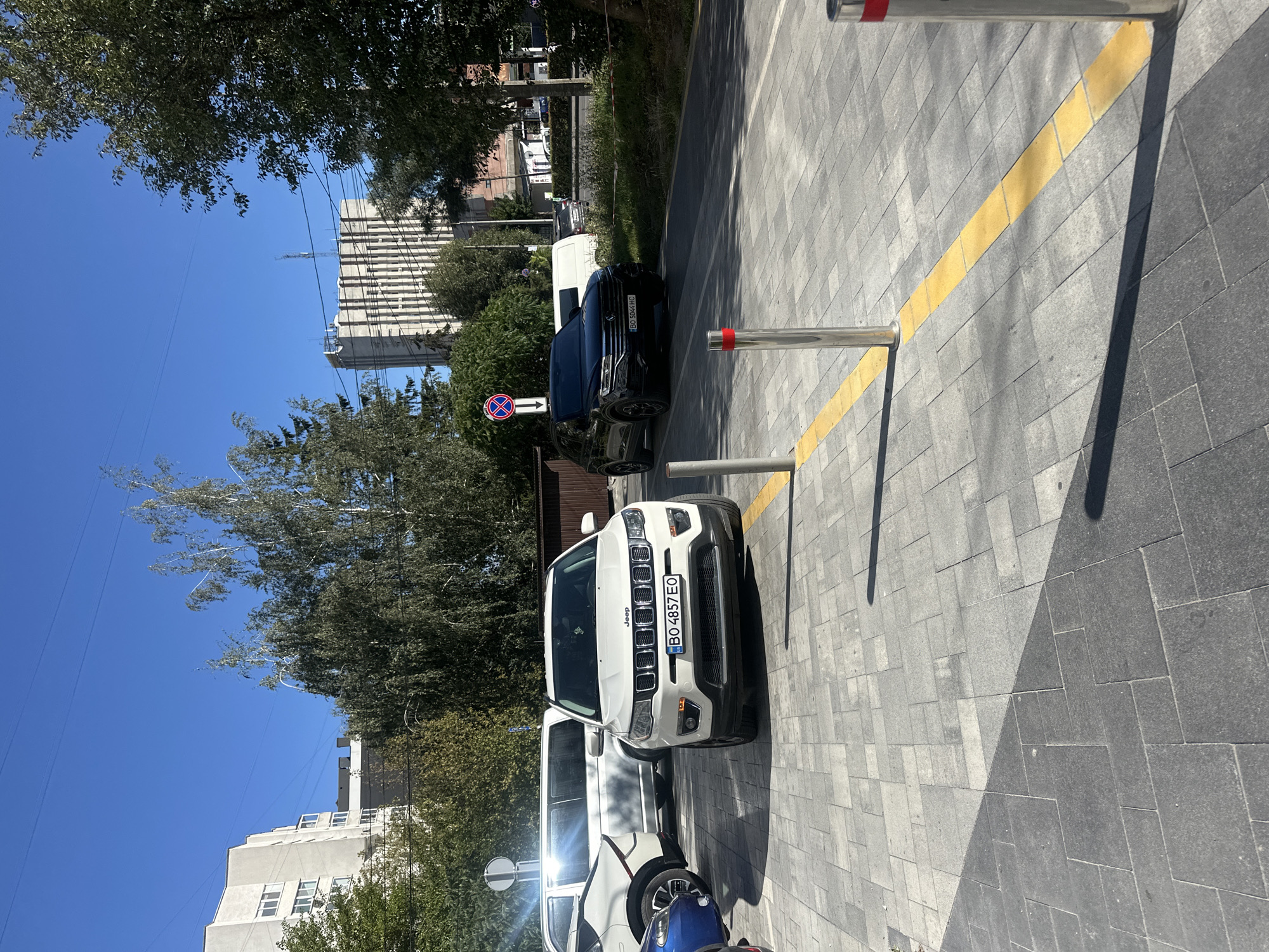 In Ternopil, Ukraine, on a clear day, a white Jeep Compass is parked on a paved area in front of buildings. The Jeep has a license plate reading "BO 4857 EO". To the right of the Jeep is a dark-colored SUV, possibly a Volkswagen Tiguan, also parked. Behind them, a white van is visible. The paving stones are gray and arranged in a rectangular pattern, with a yellow tactile paving strip running along the edge of the sidewalk. In the background, there are trees and tall buildings, including a light-colored, multi-story structure with vertical lines and a darker, more modern building. The sky is a clear blue.