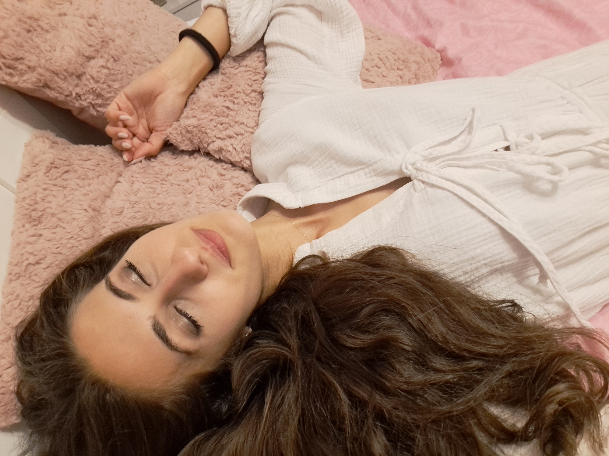 The image depicts a woman lying on her back with her eyes closed, appearing to be resting or asleep, in the city of Unknown, Unknown. Her long, wavy, dark brown hair cascades to the left side of the frame. She wears a white, textured garment, possibly a top or dress, made of a fabric with a visible weave (like gauze or linen), featuring a collared neckline and a V-neck with decorative ties at the front. Her right hand, with light-colored manicured nails, rests on a fluffy, light pink pillow. A black elastic hair tie is visible on her right wrist. A thin, delicate gold chain necklace is visible around her neck. Her eyelids display a subtle application of shimmering eyeshadow. The surface beneath her is covered by a flat pink fabric, likely a sheet or blanket. Two fluffy, light pink pillows, consistent in texture, are visible: one under her head and another to her right. The background behind the pillows is dark and indistinct, suggesting an indoor environment.