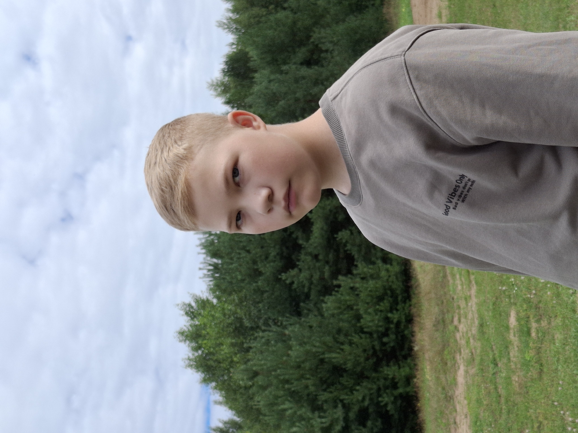 In a close-up, slightly tilted outdoor shot, a young boy with short blond hair and fair skin is the primary subject. He is looking slightly away from the camera with a neutral expression. His right ear is visible, and he is wearing a grey sweatshirt. The sweatshirt has a small, white text graphic on the sleeve that reads: "Good Vibes Only" above "Bad vibes don't go With my smile". The background consists of lush green trees and a sky with scattered clouds. The lighting suggests it is daytime, likely with overcast or partly cloudy conditions. The scene appears to be a natural setting, possibly a park or wooded area.
