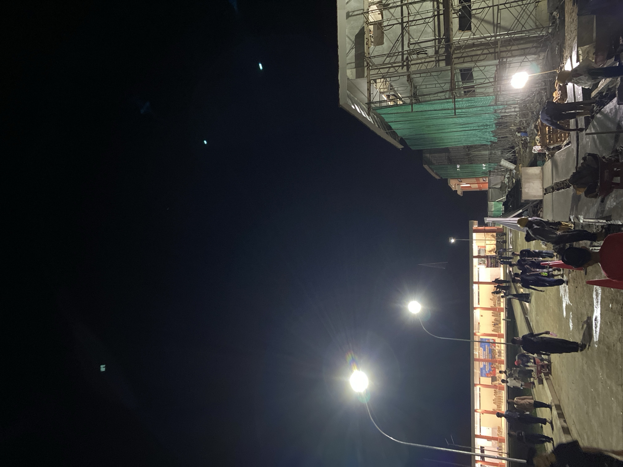 The image depicts a construction site at night in the city of Suka Makmuë, Indonesia. Several streetlights illuminate the scene, casting a bright glow on the area. In the foreground, a group of people, likely construction workers, are engaged in various activities, some appearing to be laying concrete, others carrying materials, and some walking. Some individuals are wearing hard hats and work boots. The construction site features scaffolding and green netting covering parts of unfinished buildings. In the background, a long, multi-story building with windows is visible, also illuminated. The sky above is dark, with a few faint lights or objects visible, possibly distant aircraft or drones. The overall environment suggests ongoing construction work after dark.