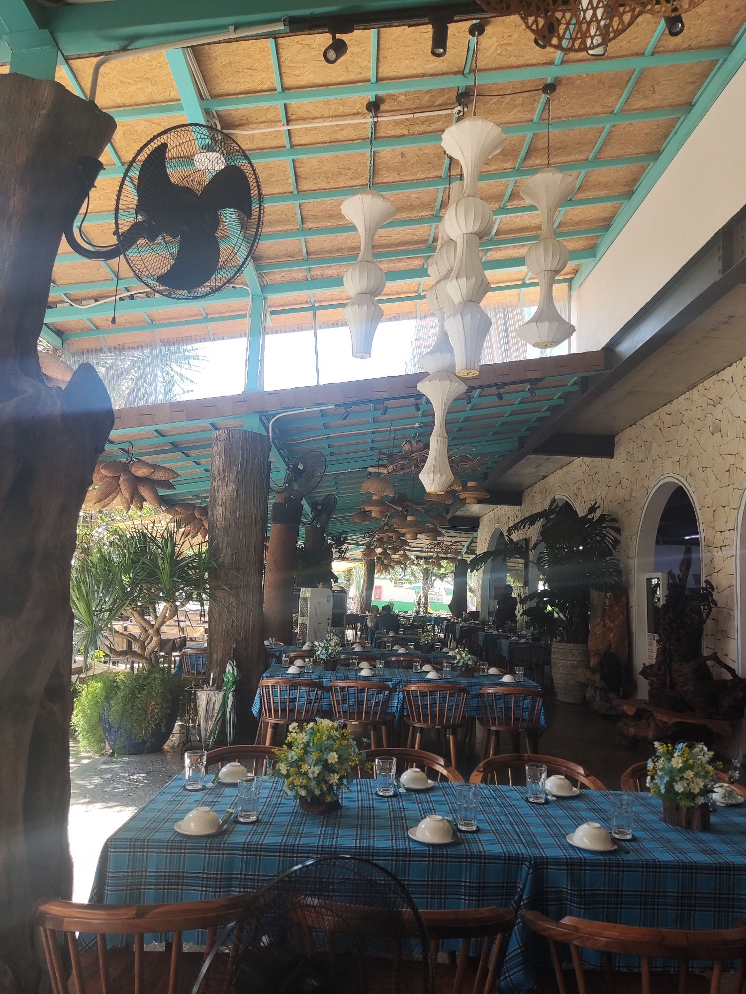 This media file captures the inviting interior of a spacious, semi-open-air restaurant or dining area, characteristic of a tropical or resort setting, likely in Đà Nẵng, Vietnam. The scene is brightly lit by natural sunlight, indicating it is daytime.
The restaurant features numerous dining tables, each meticulously set with crisp blue and white plaid tablecloths. On each table, white ceramic bowls are inverted, ready for use, alongside clear drinking glasses. Small, rustic floral arrangements, composed of white and yellow flowers, serve as charming centerpieces. The seating consists of traditional wooden chairs with slatted backs and rounded seats.
The ceiling structure is a notable feature, with a light blue-green framework supporting lighter, possibly wooden or composite, panels. Several large, industrial-style black wall-mounted fans are strategically placed throughout the space to provide cooling. Unique white pendant lights, designed with stacked, elongated bell or vase-like shapes, hang from the ceiling, adding a distinctive aesthetic. Further illumination comes from black track lighting.
Large, natural wooden pillars or tree trunks are integrated into the architecture, supporting the roof and enhancing the organic feel. Decorative elements include woven baskets or traditional fish traps suspended from the ceiling. Along one side, a textured stone wall with arched openings suggests an adjoining space. Abundant tropical plants and lush greenery are placed around the dining area, creating a vibrant, outdoor-like ambiance. Views of further outdoor seating and vegetation are visible in the background.
The dining area appears ready for guests but is currently empty, with no people visible, suggesting the photo was taken before service or during a quiet moment. The overall impression is one of peaceful preparedness and a relaxed, natural dining experience. No discernible text is visible within the image.