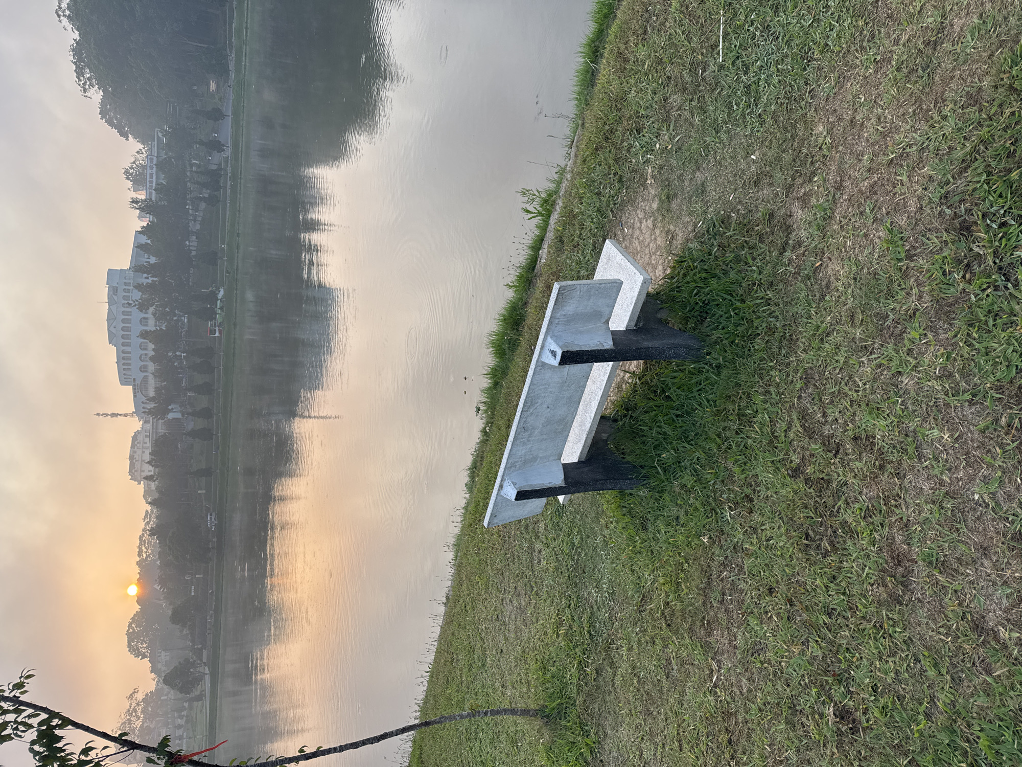 The image presents a tranquil waterside scene, likely captured during sunrise or sunset, in Da Lat, Vietnam. The perspective is from a gently sloping grassy bank overlooking a calm body of water, possibly a lake or large pond.
In the mid-ground on the right, a modern park bench constructed from light grey concrete slabs with dark metal supports sits on the grassy shore, oriented towards the water. The grass around the bench appears somewhat sparse and dry in areas, with patches of greener growth. A few bare tree branches with some leaves extend into the bottom left corner of the frame, partially framing the view.
The calm surface of the water, occupying the majority of the left side of the image, reflects the soft, warm light from the sky. Gentle ripples are visible on the water. On the distant shore, a line of trees and several buildings, including a notable large white building with arched windows, are visible through a soft haze. The sun, a bright, glowing orb, is positioned low in the sky above the distant tree line, casting a warm, diffused light across the entire landscape. The sky transitions from a soft orange-yellow near the sun to a pale grey further up, indicating either early morning or late evening.
The overall atmosphere of the scene is peaceful and serene. There is no visible text in the image.