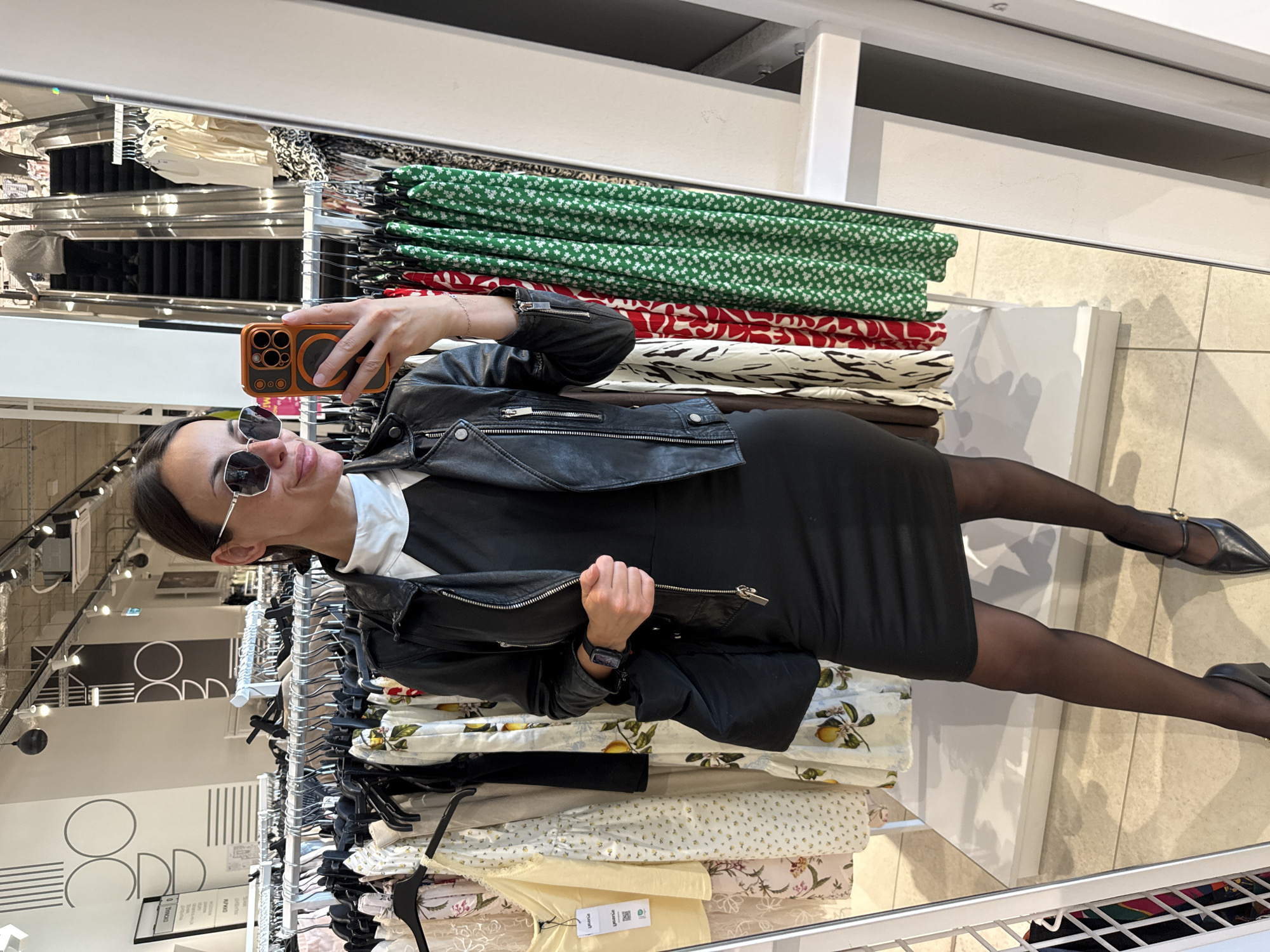 A woman wearing sunglasses, a black dress, and a black leather jacket is taking a selfie in a clothing store. She is holding an orange cell phone and standing in front of clothing racks filled with dresses and other items. She is also wearing black pantyhose and pointed shoes with straps. The environment is brightly lit and appears to be a retail setting, possibly in Pleven, Bulgaria.