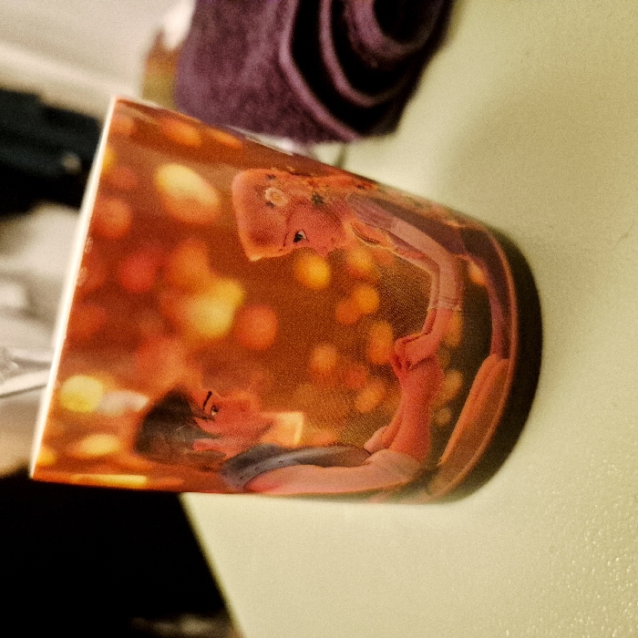 A ceramic mug with a vibrant image is presented. The design features a couple, a man lying down and a woman leaning over him, surrounded by warm orange and yellow light resembling bokeh. The style suggests a romantic or intimate scene, potentially from a movie or artwork. The colors are rich and inviting, highlighting the close interaction between the two figures. The setting appears to be a room with a textured wall.