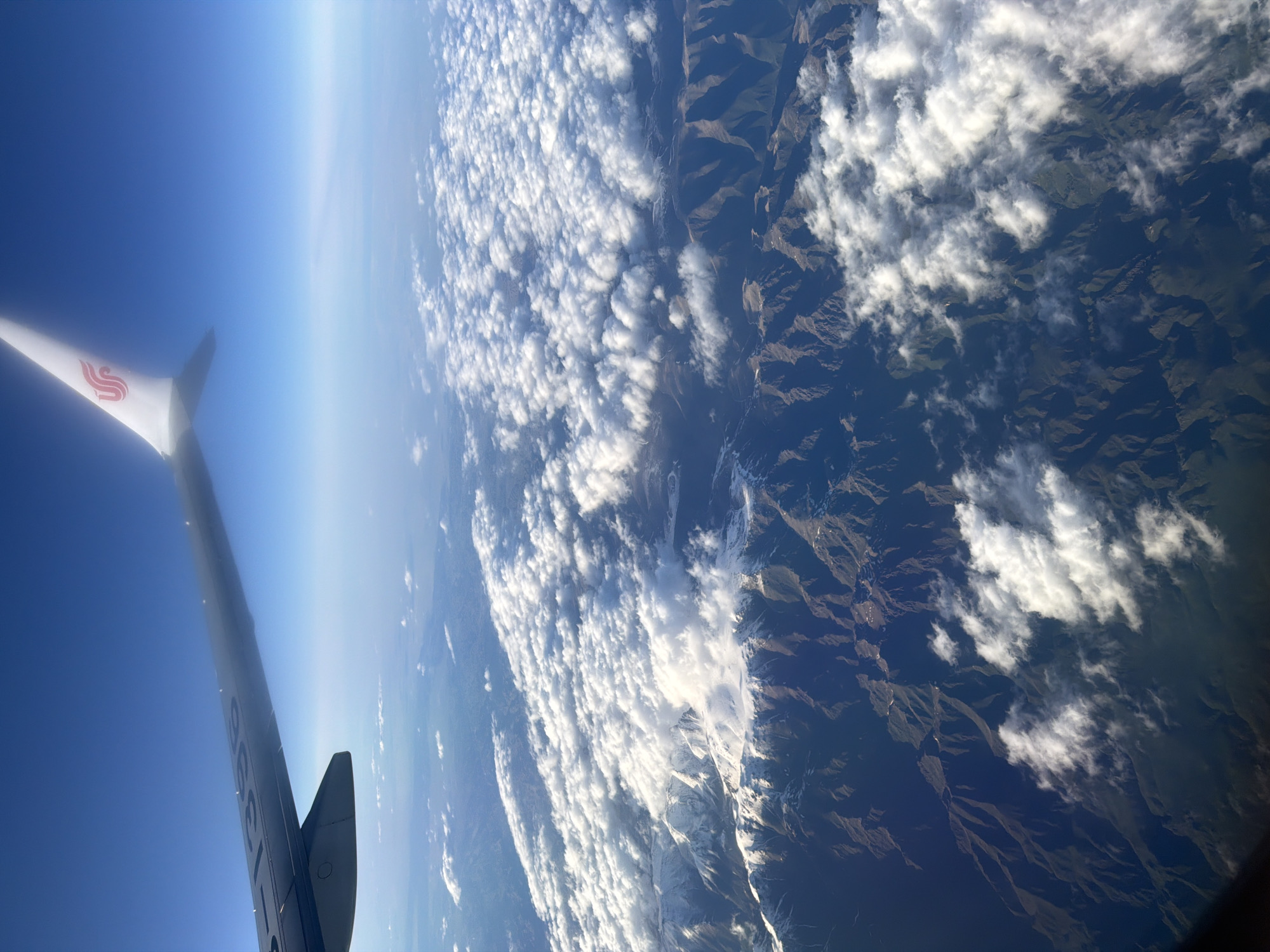 The image shows an aerial view from an airplane. A wing of the airplane, bearing the Air China logo, is visible in the left portion of the frame. The earth below is partially obscured by clouds, but mountainous terrain is visible. A thin blue atmosphere and the curvature of the earth can be seen along the horizon. The location is not the city of Khimki, Russia.