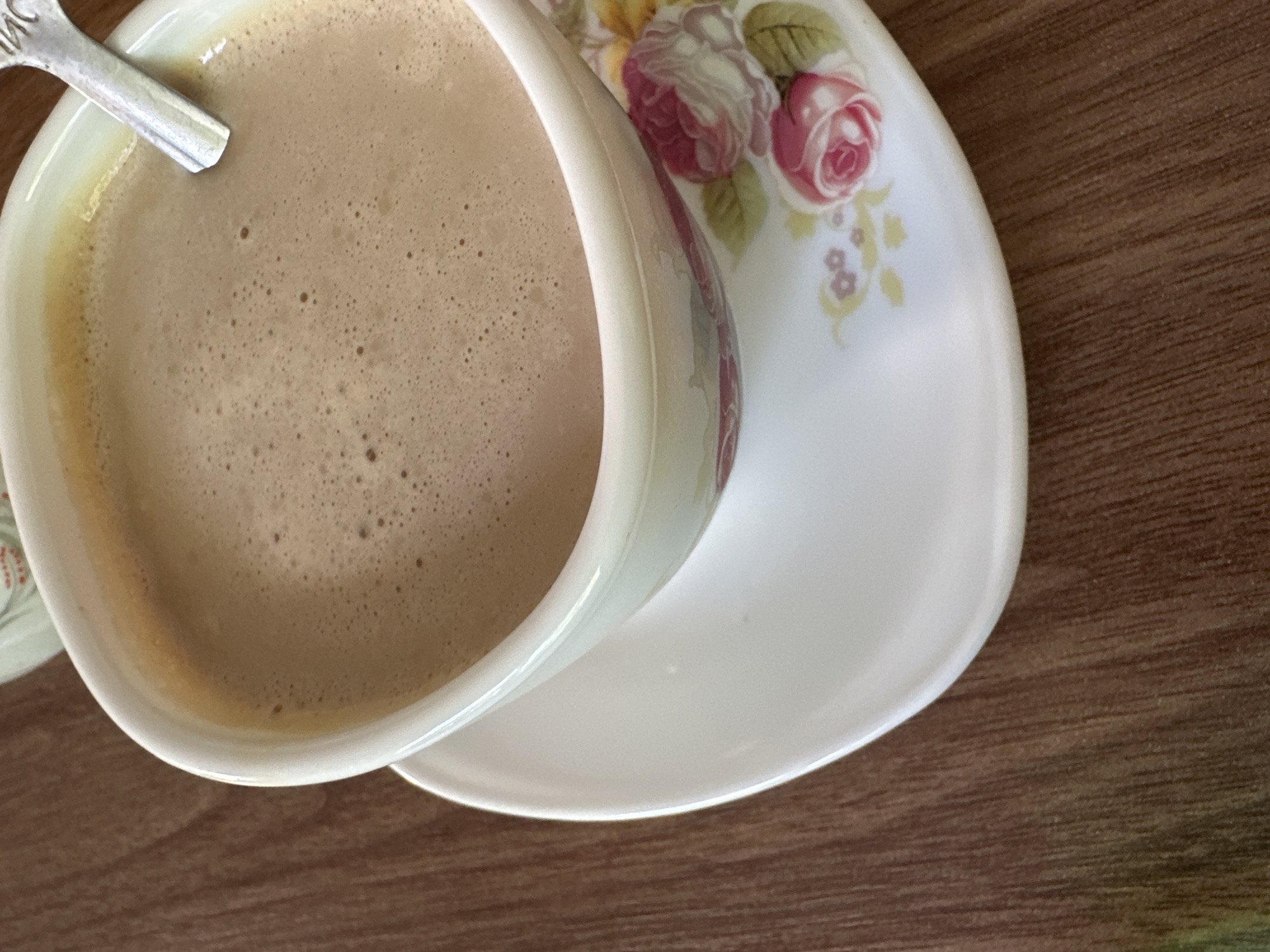 A high-angle shot depicts a cup of coffee on a saucer, resting on a wooden surface. The coffee fills most of the cup's visible interior, its surface a uniform light brown with a frothy texture. A silver spoon rests in the coffee. The cup and saucer have a white base, with floral patterns featuring roses and green foliage. The wooden surface appears to be a table. The location is unspecified in the image.