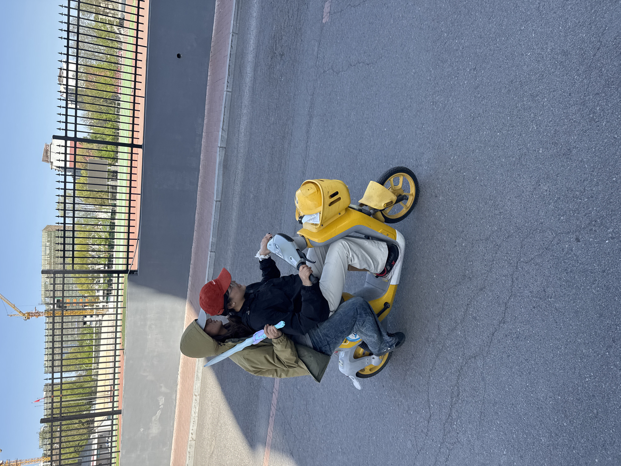Two individuals are situated on a yellow and gray e-bike on an asphalt surface. One individual wears a red baseball cap, a black jacket, and blue jeans. The other individual wears a tan hooded jacket. The individual in the tan jacket is holding a light blue toy sword. Behind them, there's a black metal fence adjacent to a gray wall. Across the fence, a sports field and buildings under construction are visible, including a yellow crane. The image was taken during daylight hours. Location: Zhongtun, China.