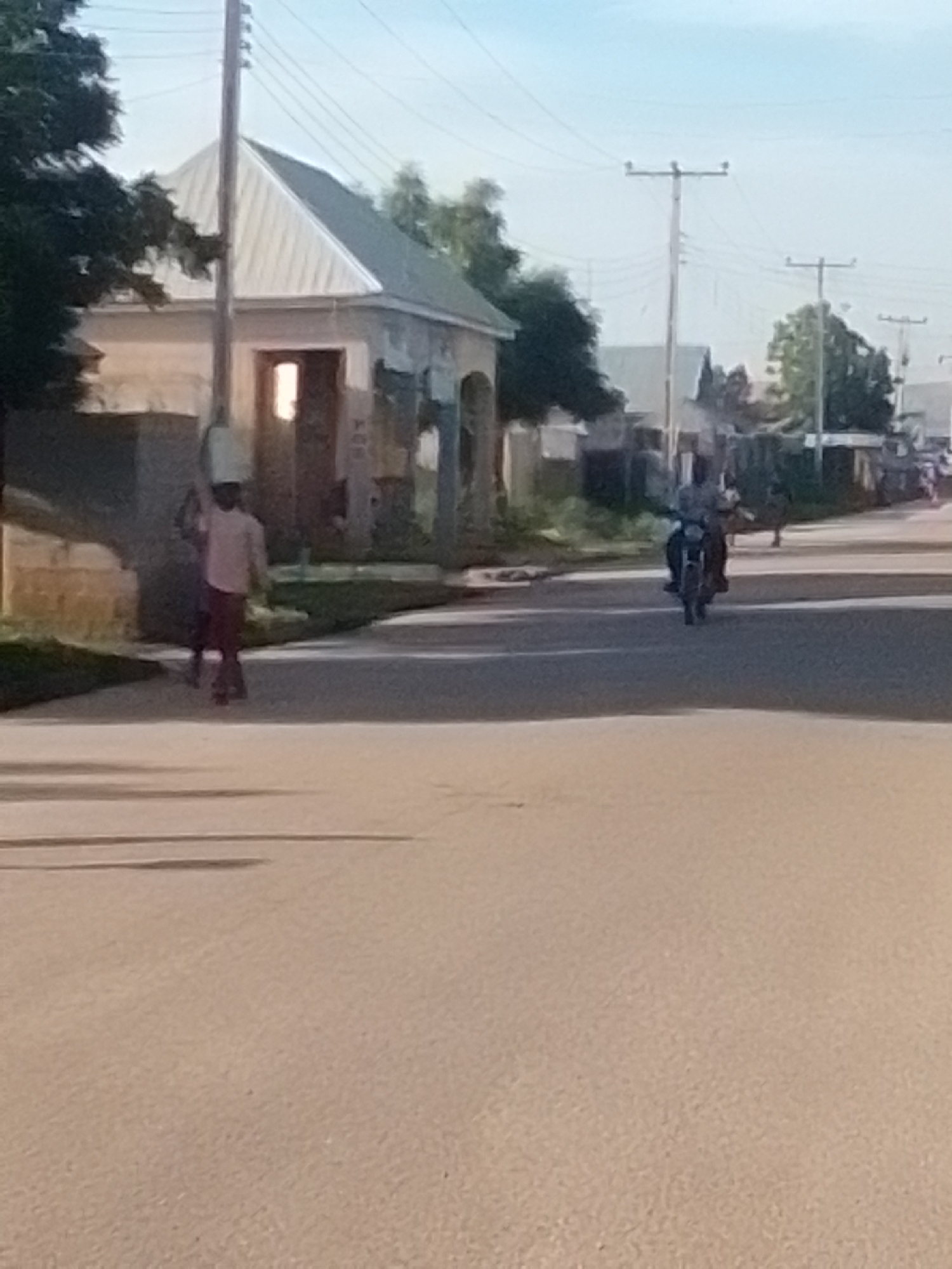 The image depicts a street scene in Liji Hill, Nigeria. On the left side of the paved road, a young person is walking with a white bucket balanced on their head. The person is wearing a light-colored long-sleeved shirt and dark pants. Behind them, a building with a white, peaked roof and light-colored walls is visible. A utility pole with wires runs vertically in front of the building. Further down the street, a motorcycle is being ridden by a person, moving away from the camera. The environment includes more buildings and trees in the background. The sky is a pale blue. The lighting suggests it is daytime, possibly late afternoon.