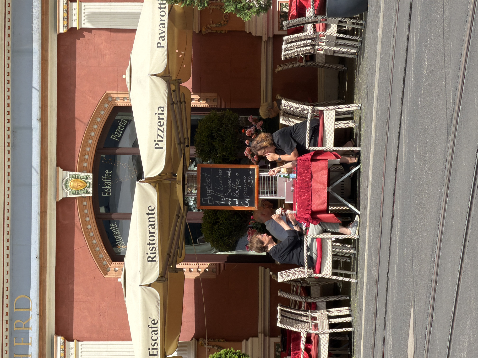 The image depicts an outdoor seating area of a restaurant in Erfurt, Germany, indicated by the word "ERFURT" visible on the building's facade. Two women are seated at a small table covered with a red tablecloth. The woman on the left, wearing a black top and grey pants, is eating an ice cream. The woman on the right, also in a black top and grey pants, is looking at her phone. Another person, partially visible, is seated behind them. Yellow umbrellas with the words "Eiscafe", "Ristorante", and "Pizzeria" are extended over the seating area. A chalkboard menu is displayed on an easel. In the background, there is a red building with architectural details and windows. The seating area is adjacent to a street with tram tracks. The overall scene is that of a sunny day at a European cafe.